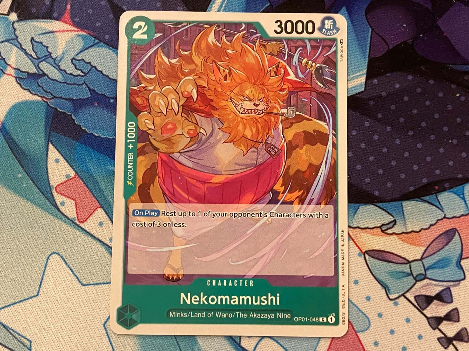 Nekomamushi 桌布 壁紙 — 一張貓蝮蛇的火熱動漫卡牌，放置在帶有蝴蝶結和星星的彩色圖案布料上。