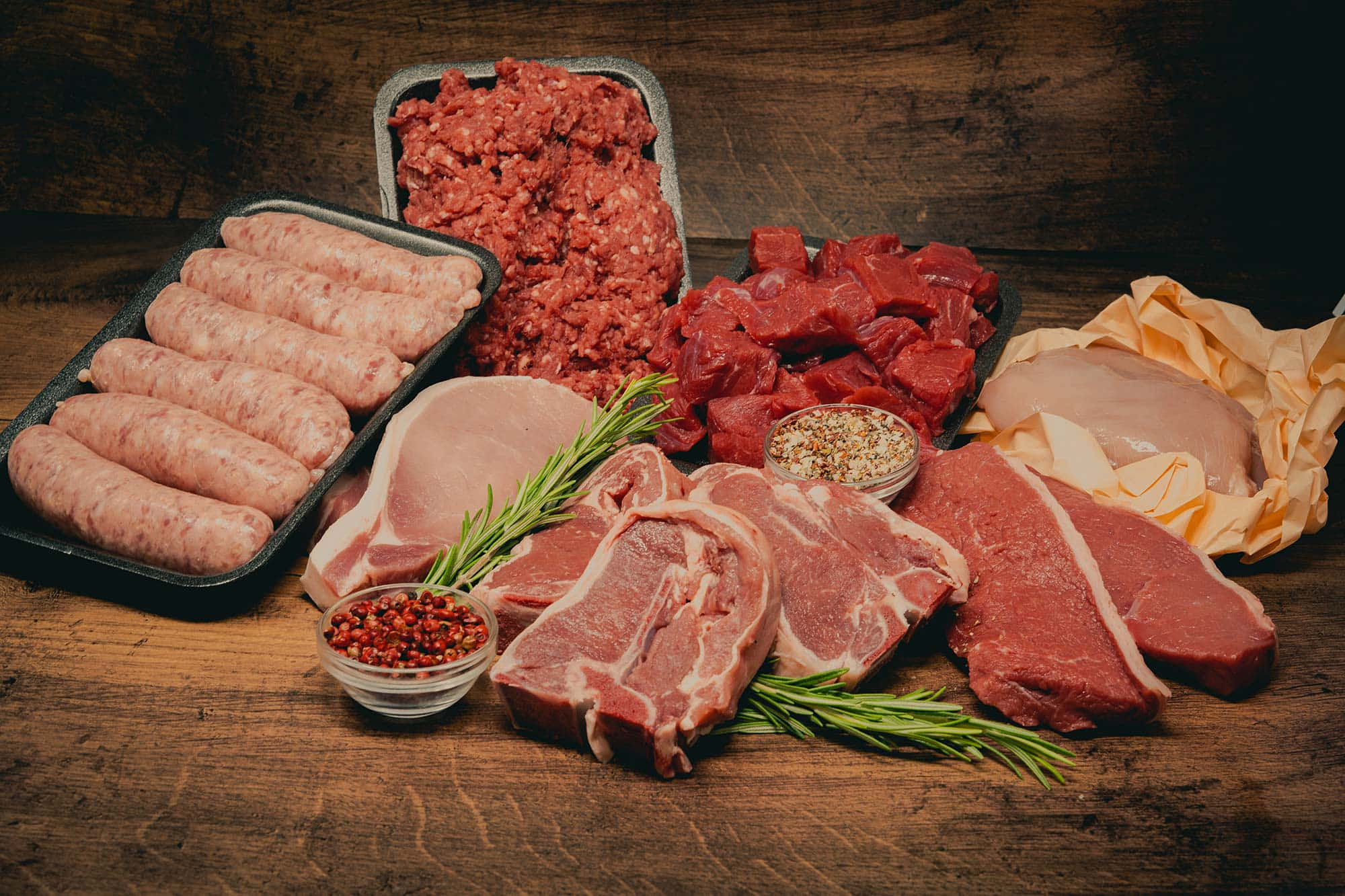 meat 桌布 壁紙 — 一張質樸的木桌上擺放著各種生肉，包括香腸、碎牛肉、牛扒、豬扒和雞胸肉，並以迷迭香和胡椒粒裝飾。