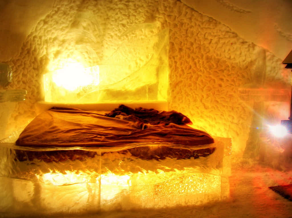 Ice Hotel 桌布 壁紙 — 一張鋪著深色布料的舒適床鋪，安放在一個溫暖照明的冰洞內，洞壁和天花板散發著琥珀色的光芒。