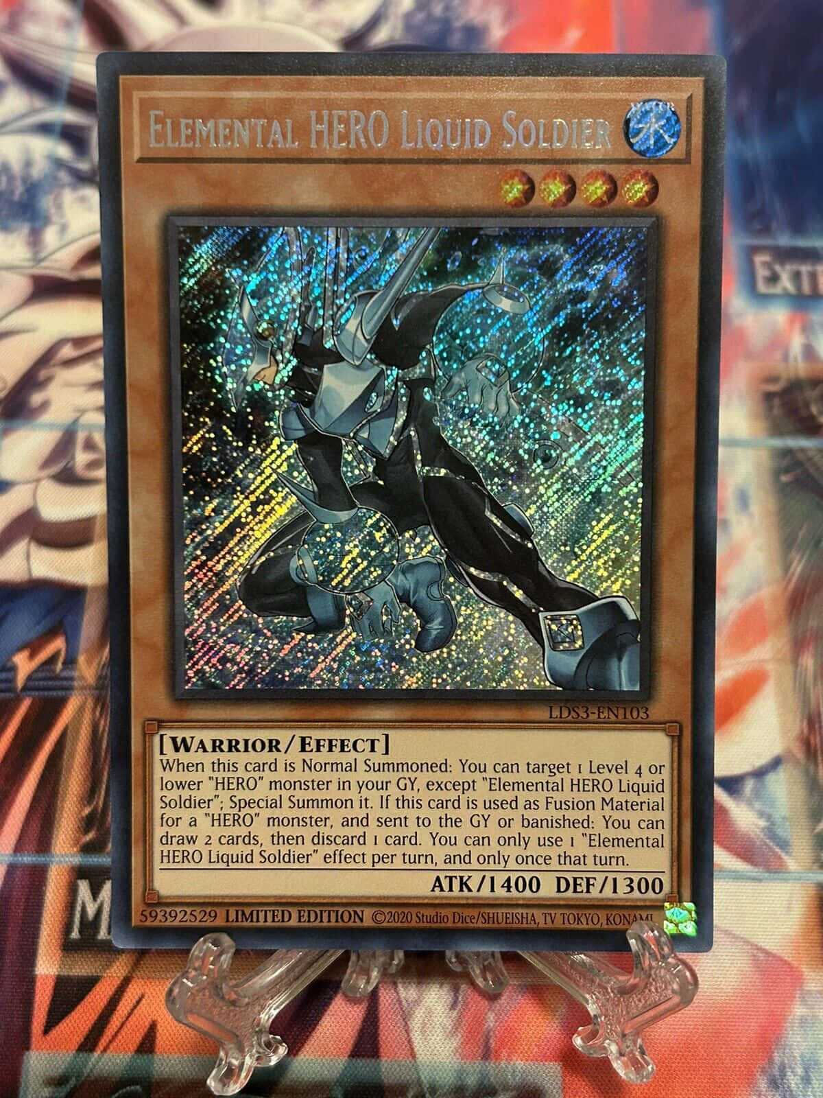 Yugioh Elemental Hero 桌布 壁紙 — 一張限量的遊戲王卡牌元素英雄液體士兵立在透明支架上，背景是色彩繽紛的動漫主題。