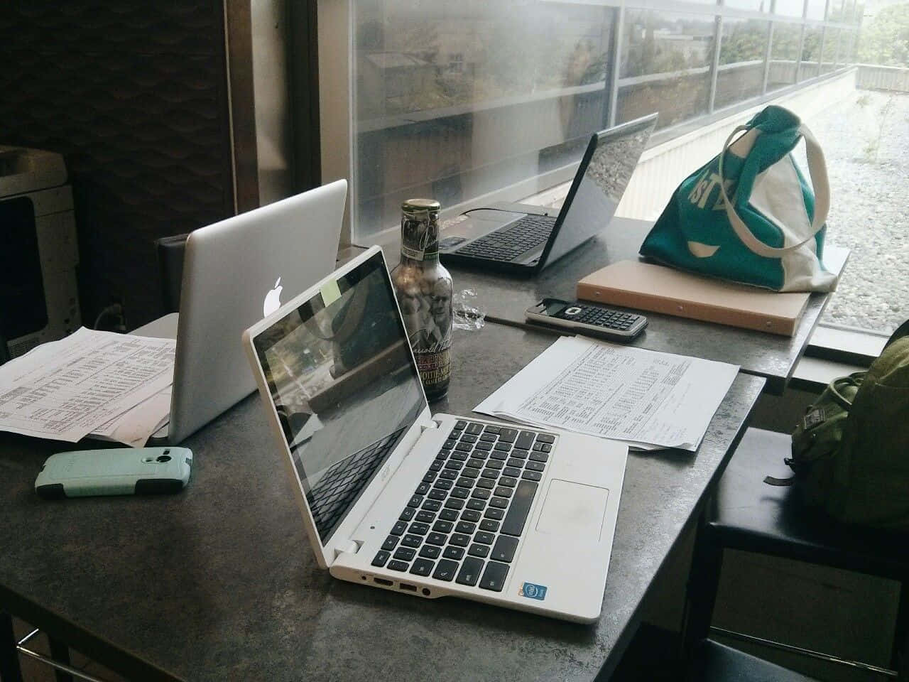 溫習 桌布 壁紙 — 一張雜亂的書桌，上面有兩部打開的筆記型電腦、文件、一個瓶子和一個袋子，靠近一扇顯示下雨戶外場景的窗戶。