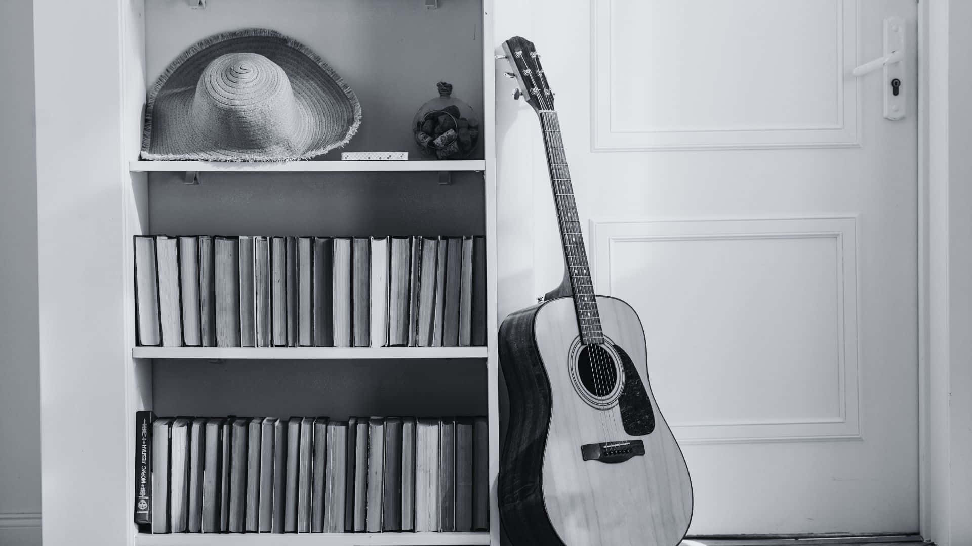 Black And White Guitar 桌布 壁紙 — 一張黑白照片，一把原聲結他靠喺一個放滿書本同草帽嘅書架旁邊。