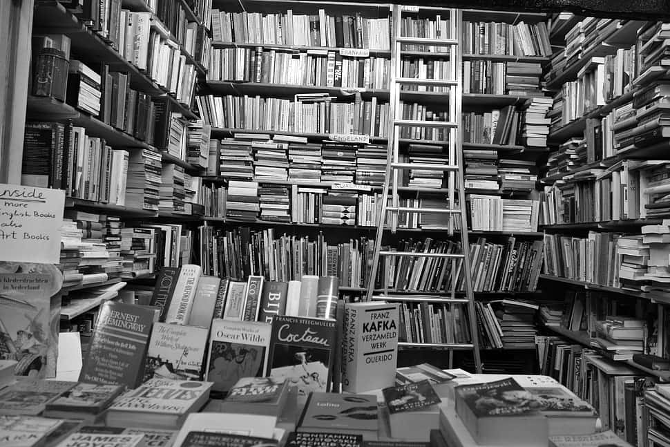 Black And White Book 桌布 壁紙 — 一張黑白照片，展示了一間雜亂的書店，有高聳的書架、梯子，以及可見的作者名字，如卡夫卡和王爾德。