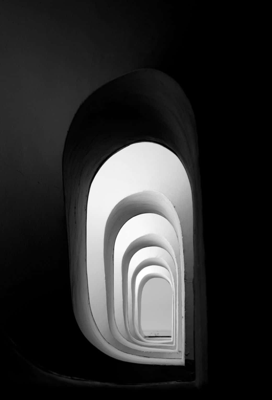 Black And White Architecture 桌布 壁紙 — 一張黑白照片，展示嵌套的拱形門廊逐漸消失在黑暗中，形成一條光之隧道。