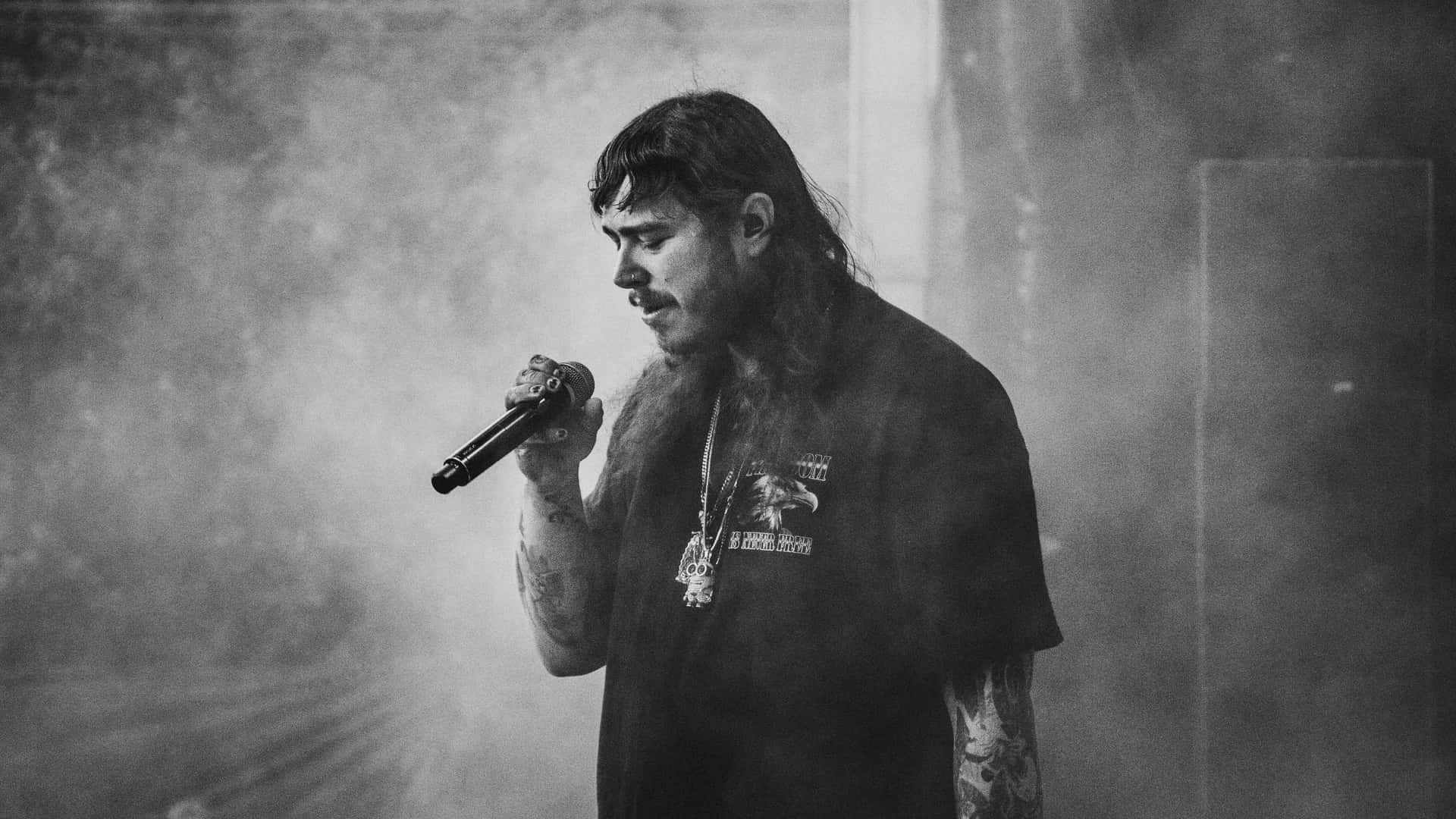 Post Malone 桌布 壁紙 — 一張黑白照片，拍攝一位紋身長髮男子在煙霧彌漫的舞台上對著咪高峰唱歌。