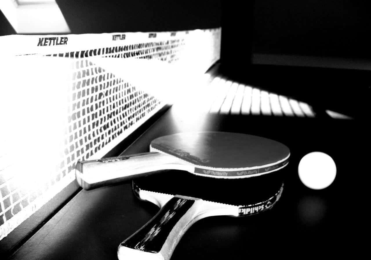 Table Tennis 桌布 壁紙 — 一張黑白照片，拍攝了在戲劇性燈光下，乒乓球拍和球放在明亮球網旁邊的桌子上。