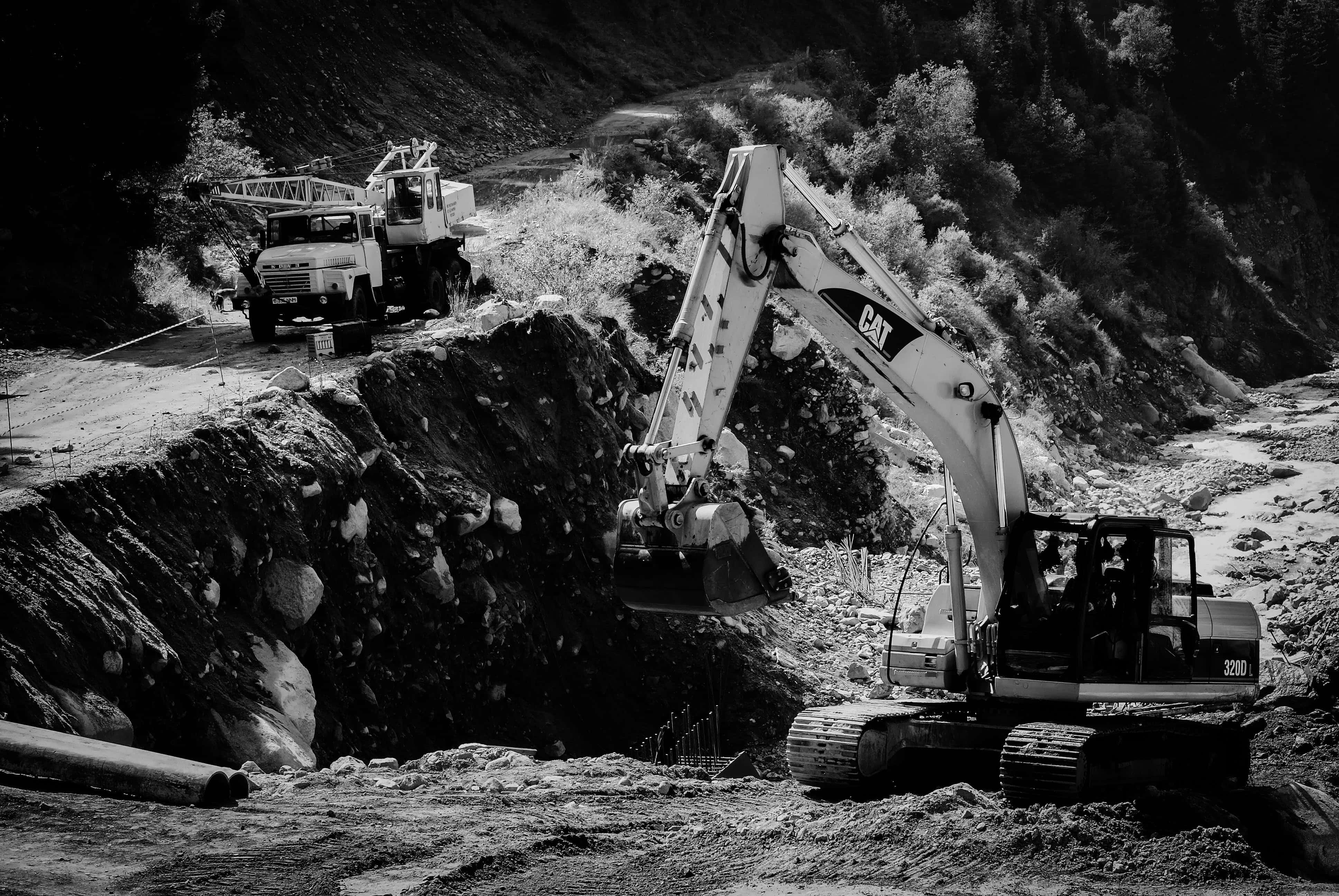 Mining 桌布 壁紙 — 一張黑白照片，顯示一輛CAT挖掘機正在停泊的工程車旁邊的岩石山坡上挖掘。