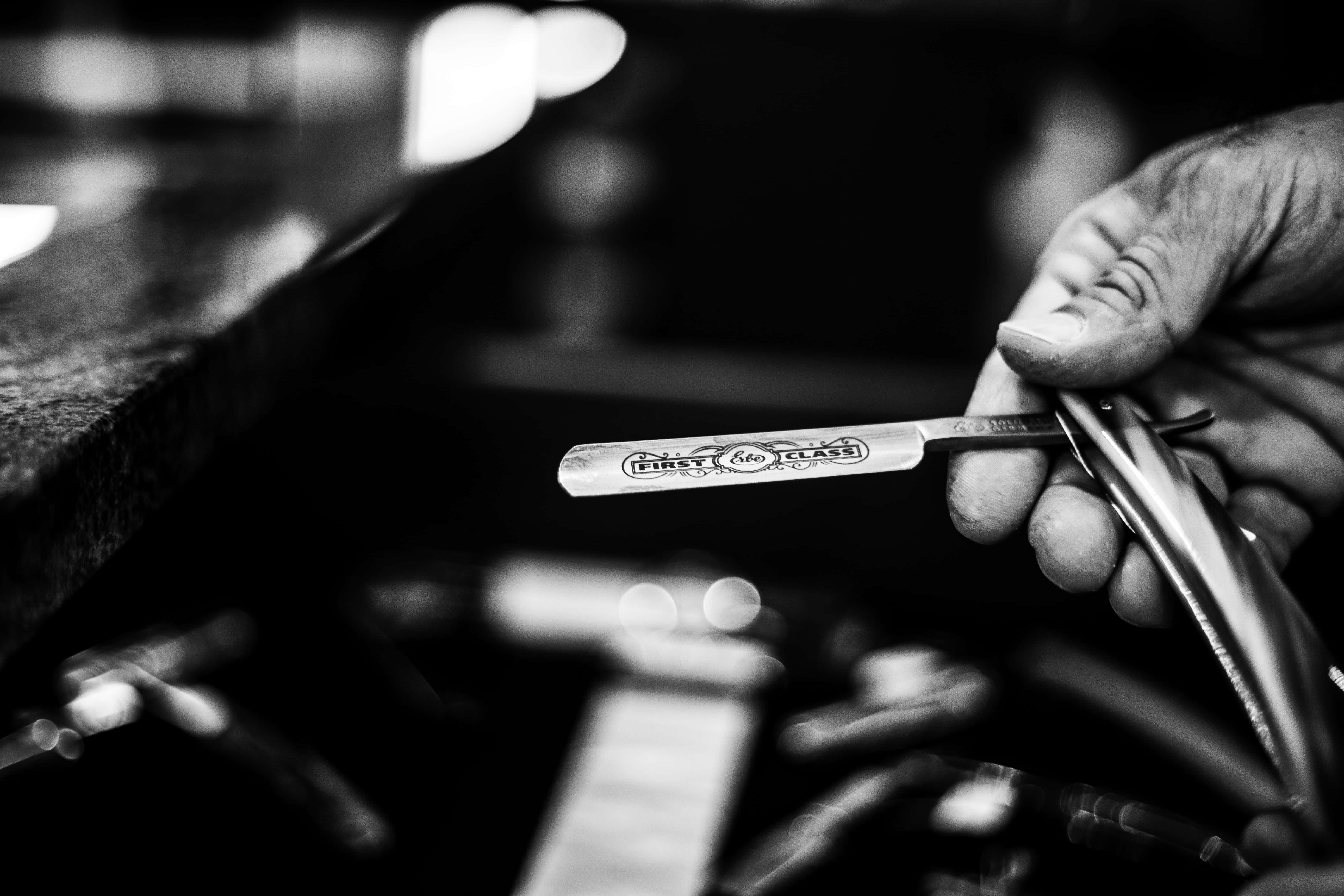 Barber 桌布 壁紙 — 一張黑白特寫，一隻手拿著刻有'FIRST CLASS'的復古直剃刀，背景是模糊的理髮店。