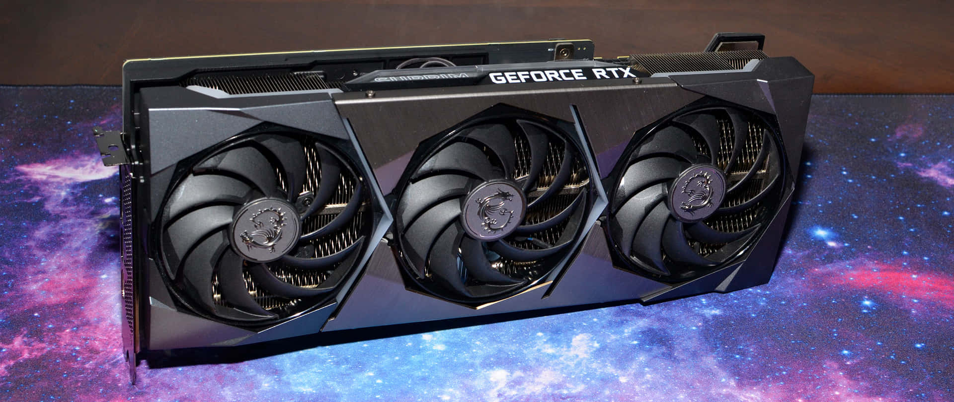  壁紙 — 一張黑色MSI GeForce RTX顯示卡，配備三個風扇，放置在一個帶有紫色、藍色和粉紅色星星的宇宙星雲壁紙上。