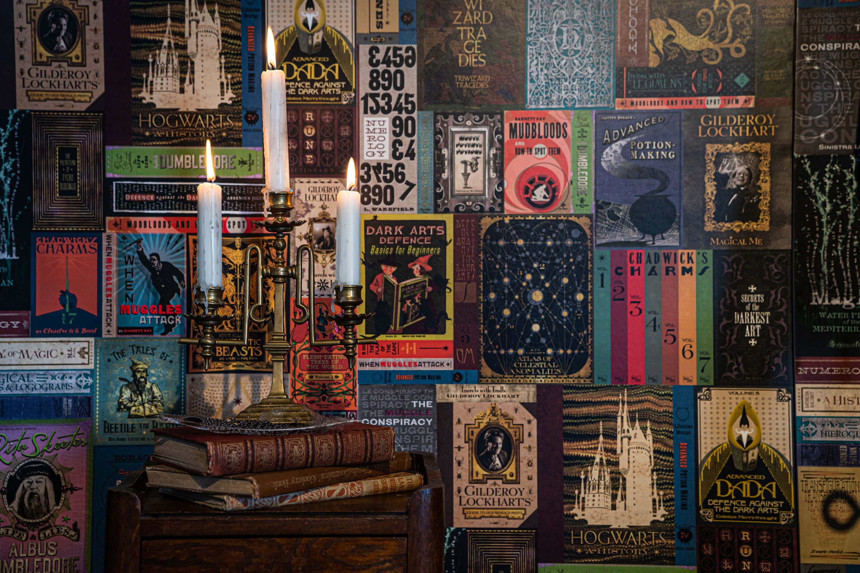The Hogwarts Library 桌布 壁紙 — 一張點著蠟燭的木桌上放著疊起的書本，前面是鋪滿復古哈利波特書封的牆壁。