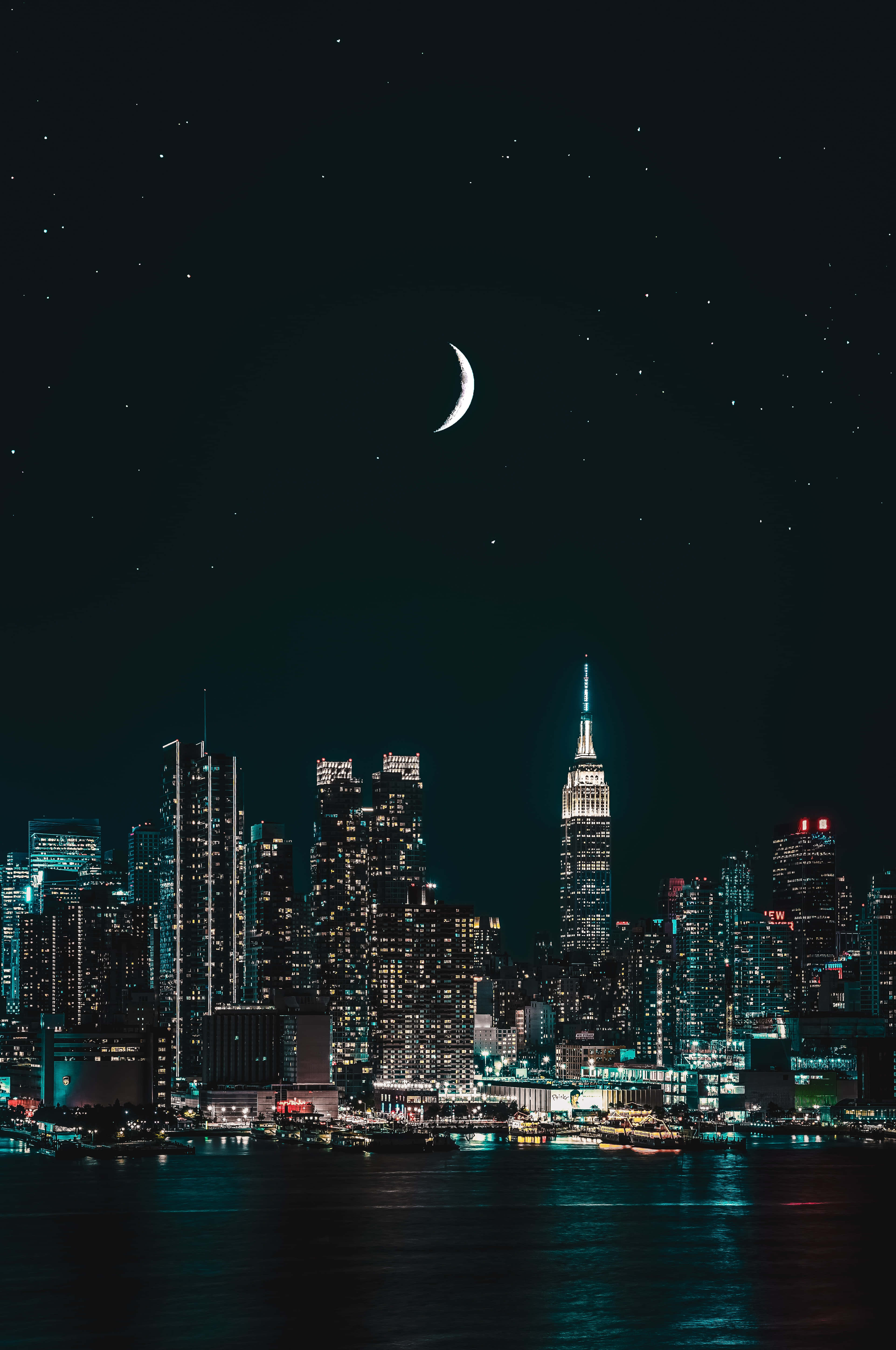 城市景觀 桌布 壁紙 — 一彎新月懸掛在閃爍的夜景城市上空，摩天大樓在星空下的暗水中倒映。