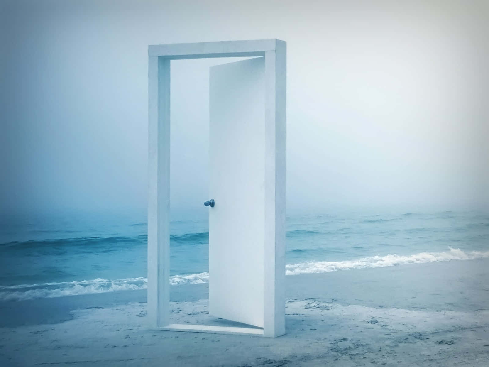 Door 桌布 壁紙 — 一扇孤獨的白色門半開在霧氣瀰漫的海灘上，露出門框外的海洋。