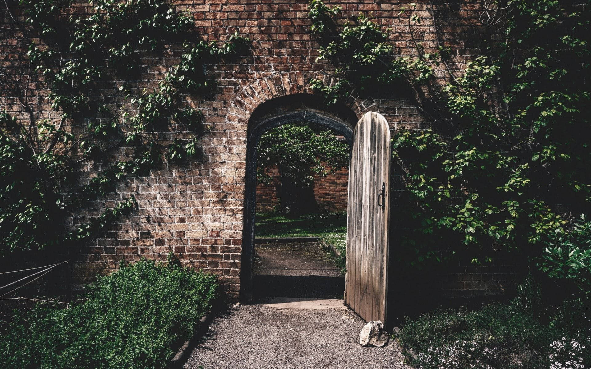 Stone Wall 桌布 壁紙 — 一扇舊木門半開在長滿青苔的磚拱門中，露出後面陽光普照的花園小徑。