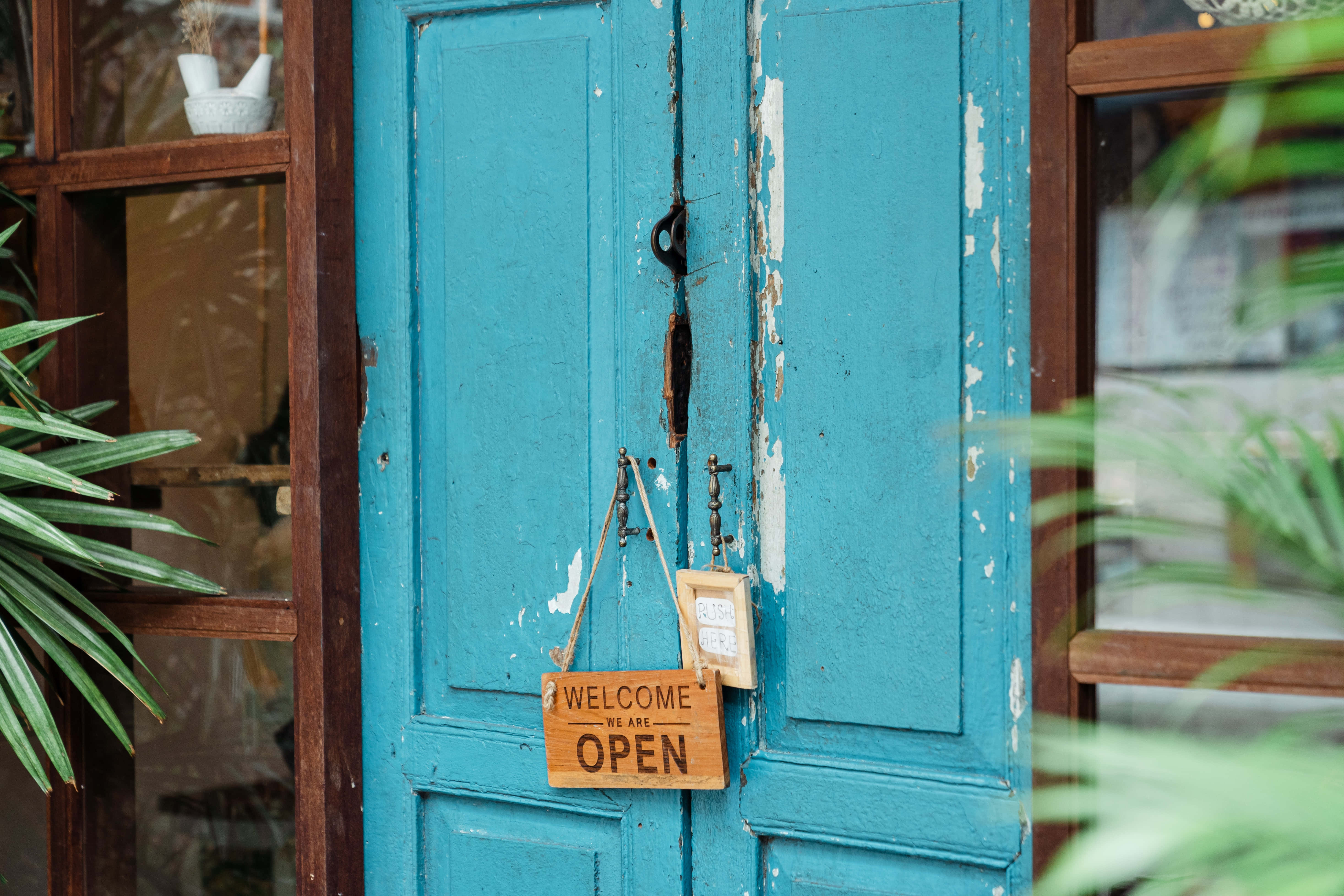 Welcome 桌布 壁紙 — 一扇風化的藍綠色門，油漆剝落，木製的「WELCOME WE ARE OPEN」標誌懸掛在玻璃窗之間，周圍環繞著茂盛的綠色植物。