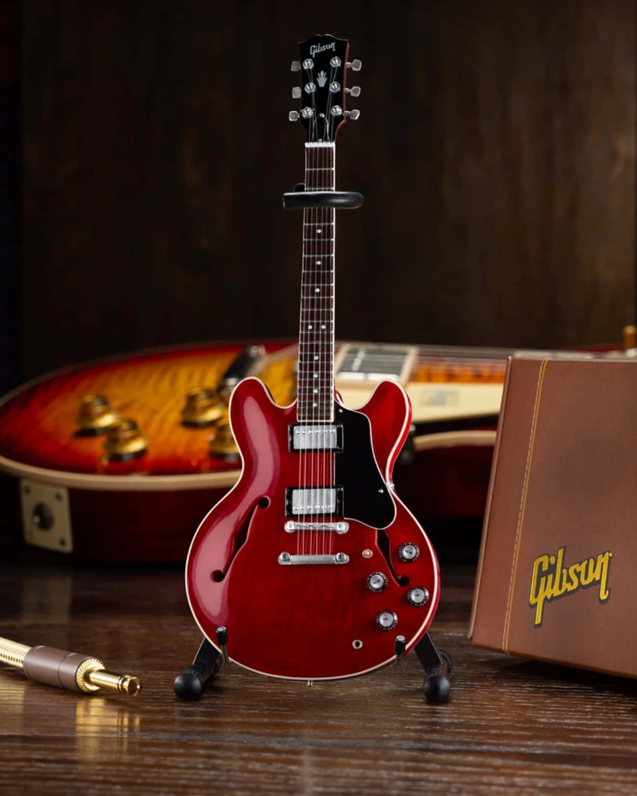 Axe Heaven Gibson 335 壁紙 — 一把光澤的紅色Gibson結他直立於木質表面上，旁邊有一個復古結他盒和一條線纜。