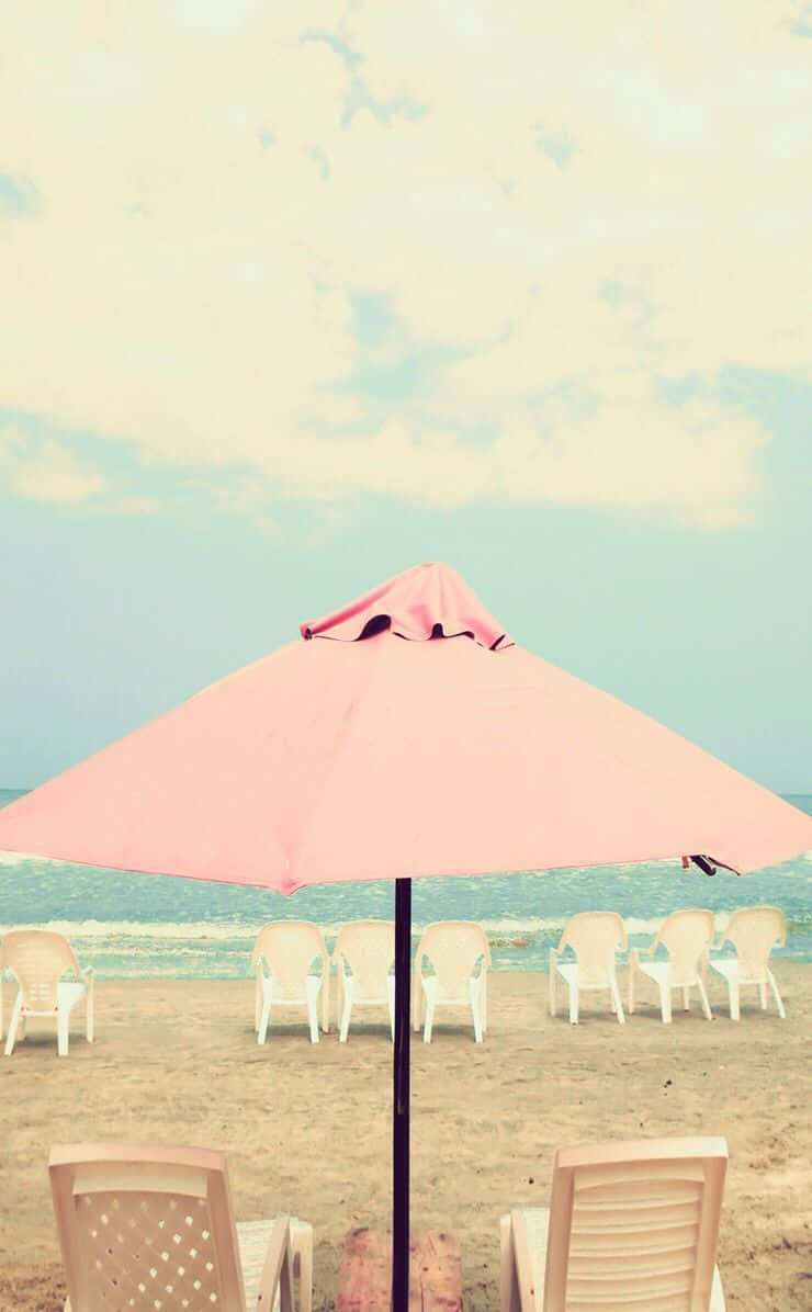 Beach Umbrella 桌布 壁紙 — 一把柔和的粉紅色沙灘傘遮擋著沙灘上的白色椅子，伴隨著輕柔的海浪和多雲的天空。