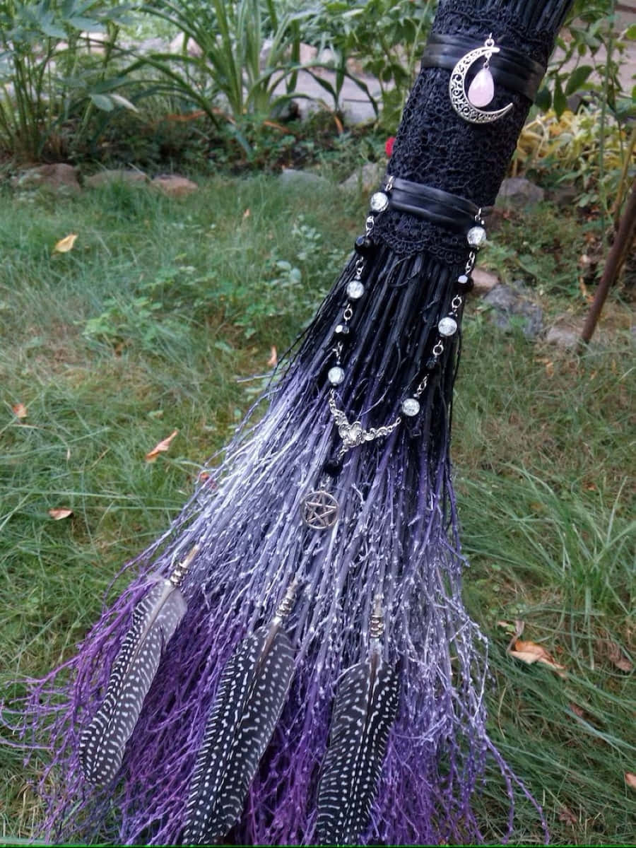 Witches Broom 桌布 壁紙 — 一把神秘的女巫掃帚，帶有黑色蕾絲手柄、月亮吊墜、紫色羽毛和銀色飾物，躺在長滿草的花園中。
