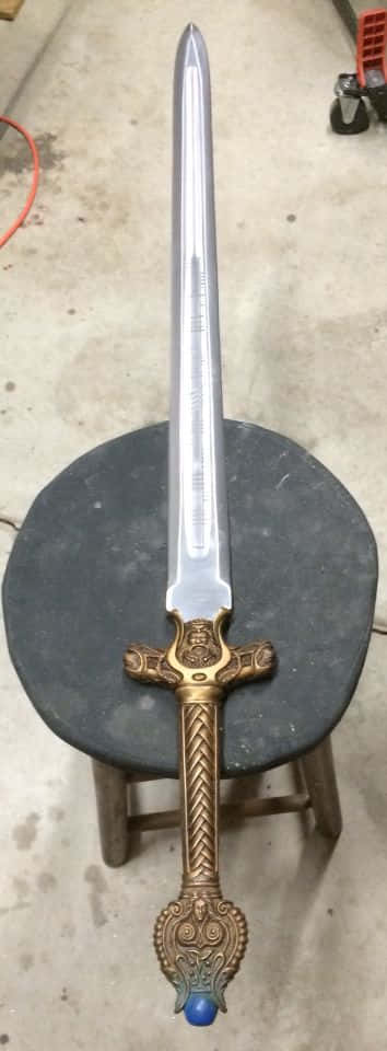 Excalibur 桌布 壁紙 — 一把細節豐富的裝飾劍，金色劍柄和藍色劍首，直立放在混凝土工作室的黑色凳子上。