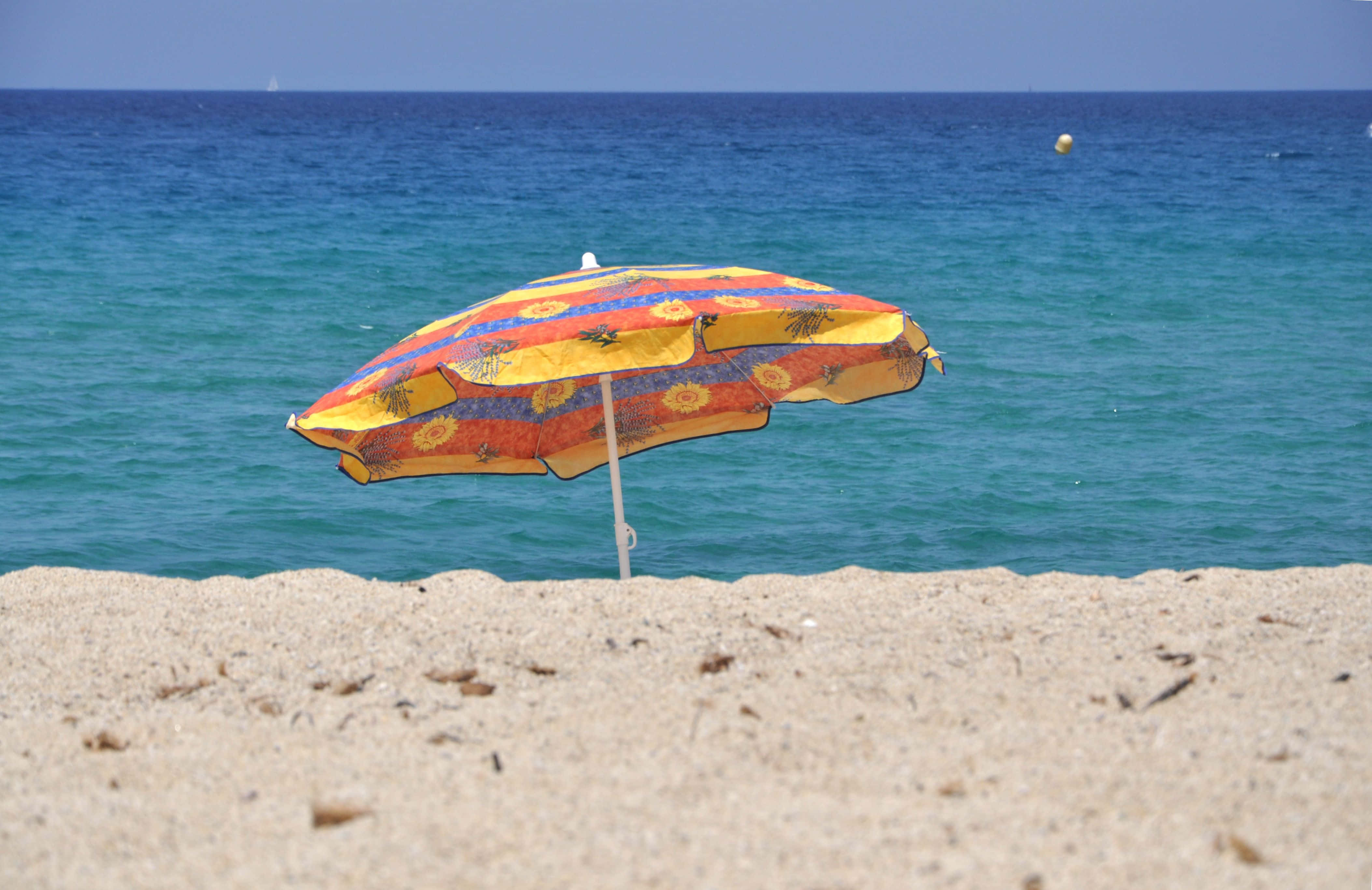 Beach Umbrella 桌布 壁紙 — 一把色彩繽紛、帶有太陽圖案的沙灘傘獨自立在沙灘上，面向碧綠色的海洋，天空清澈湛藍。