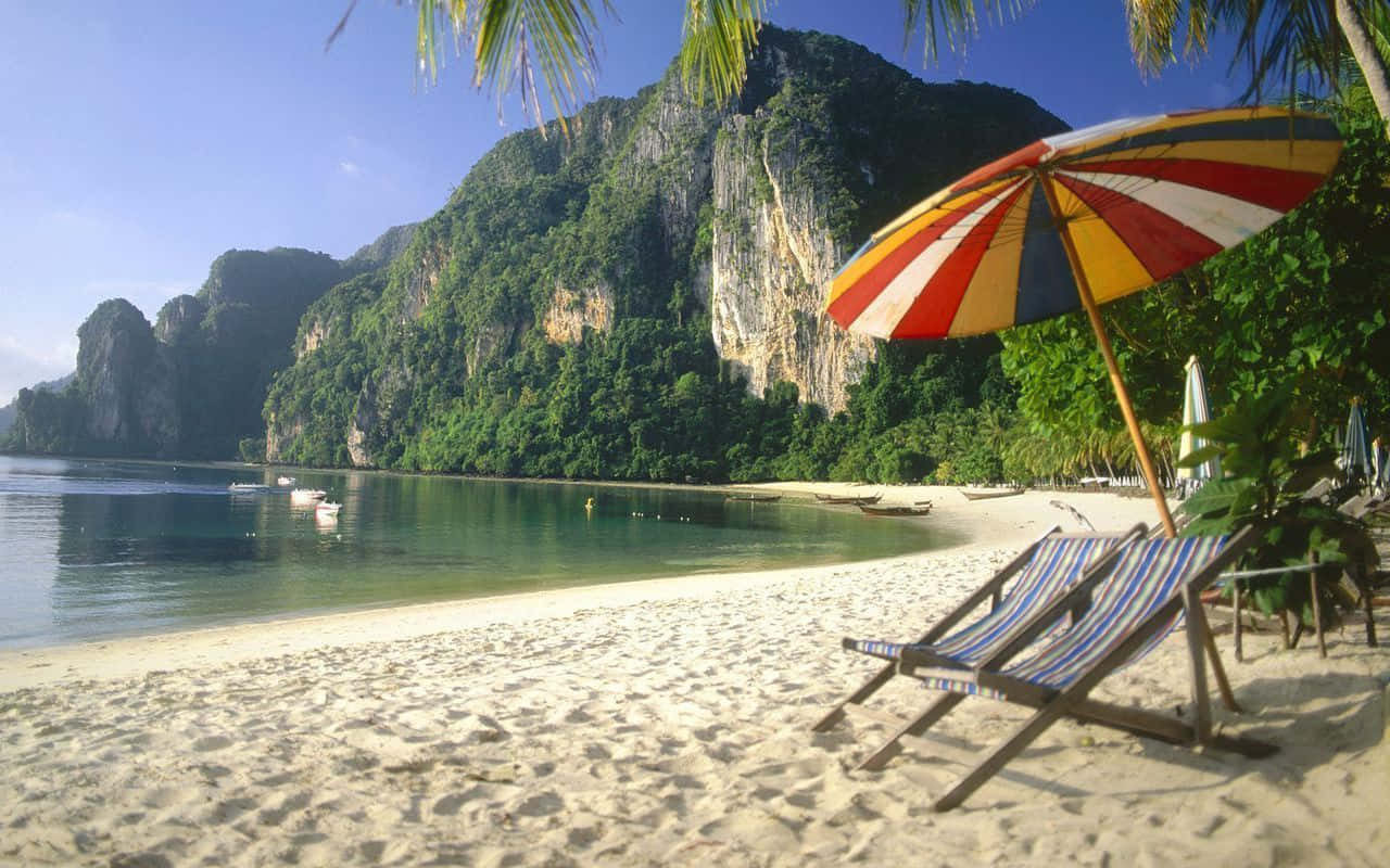 Thailand Beach 桌布 壁紙 — 一把色彩鮮豔的條紋遮陽傘在純淨的白色沙灘上為躺椅遮蔭，沙灘旁有碧綠的海水和高聳的綠色懸崖，上方是清澈的藍天。