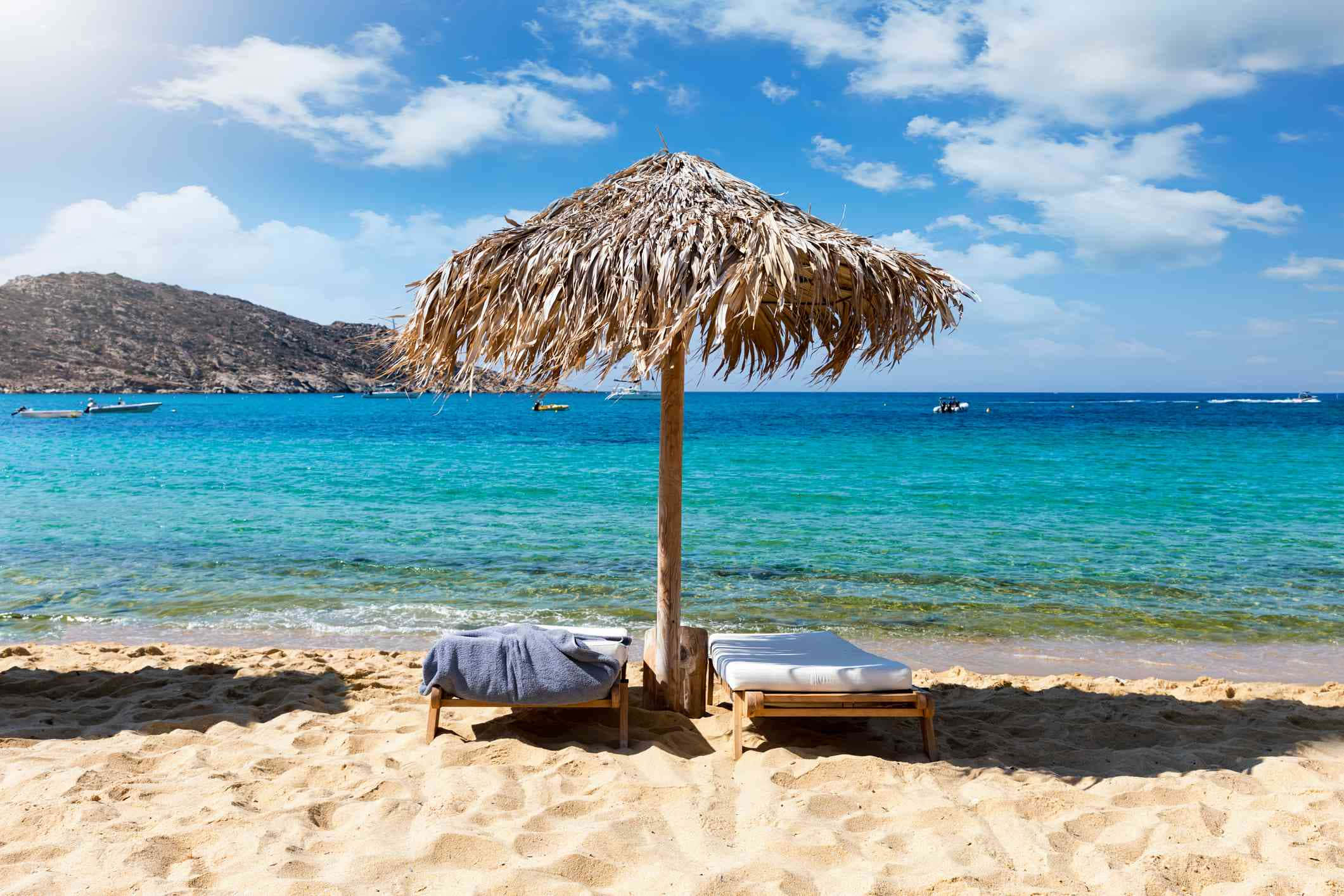Greek Beach 桌布 壁紙 — 一把茅草遮陽傘為兩個躺椅遮蔭，位於陽光明媚的沙灘上，有碧綠的海水和遠處的山丘，在明亮的藍天下。
