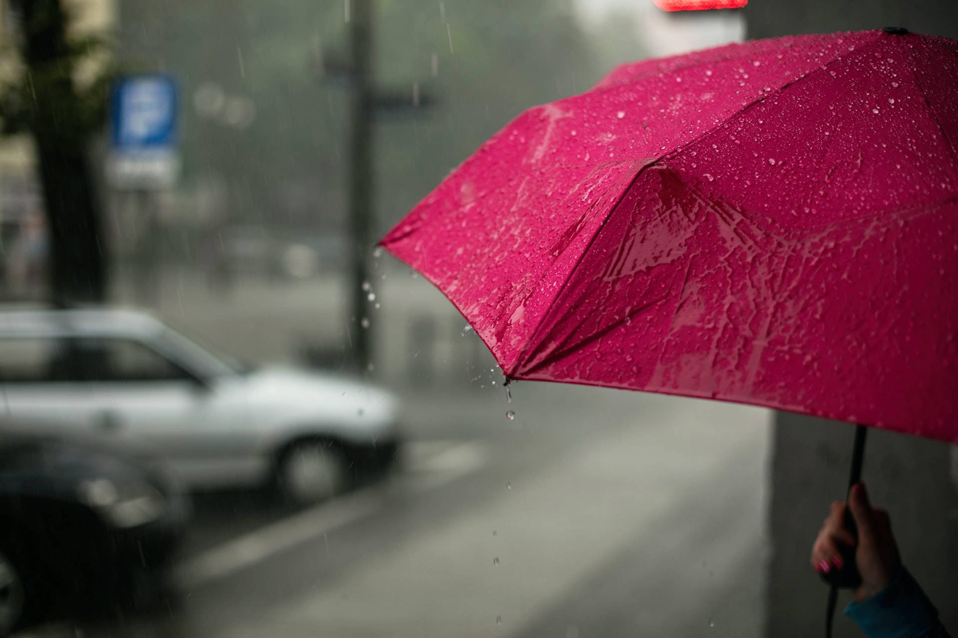 落雨 桌布 壁紙 — 一把鮮豔的粉紅色雨傘在暴雨中，水滴落在模糊的城市街景上。