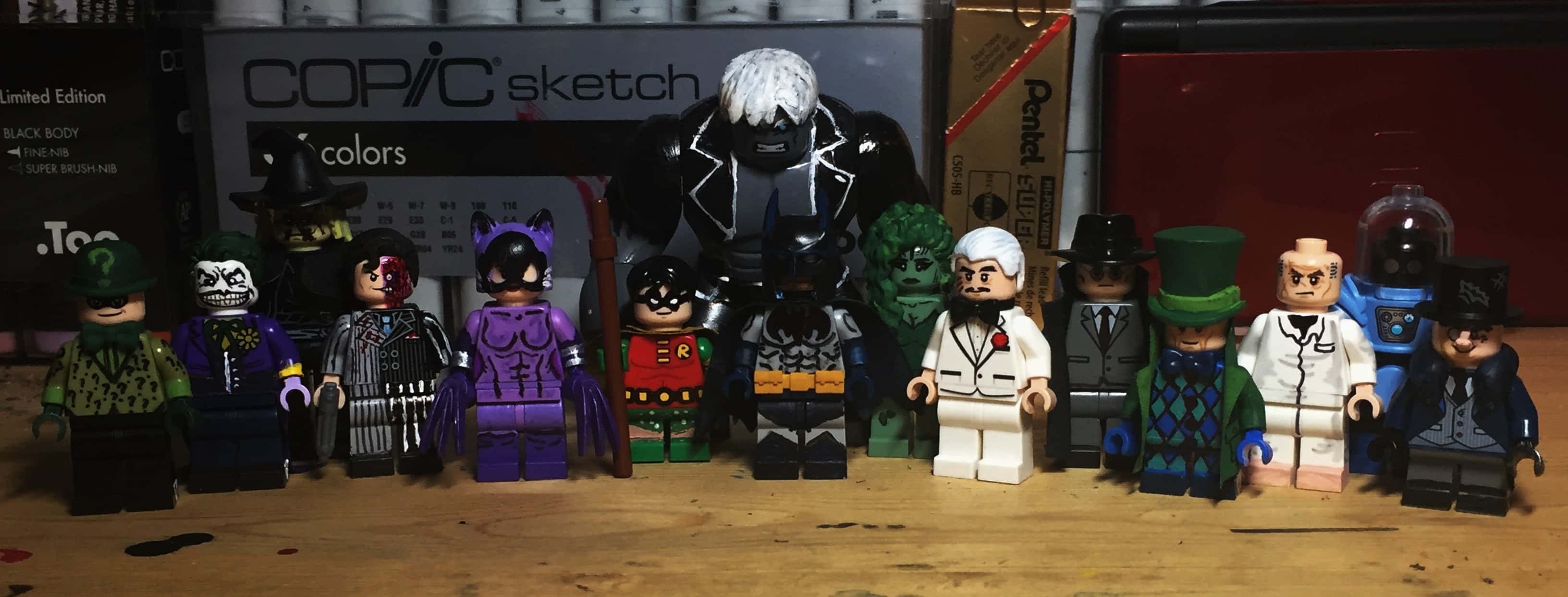 Batman Dark Victory 桌布 壁紙 — 一排LEGO蝙蝠俠反派在木桌上擺姿勢，背景有美術用品。