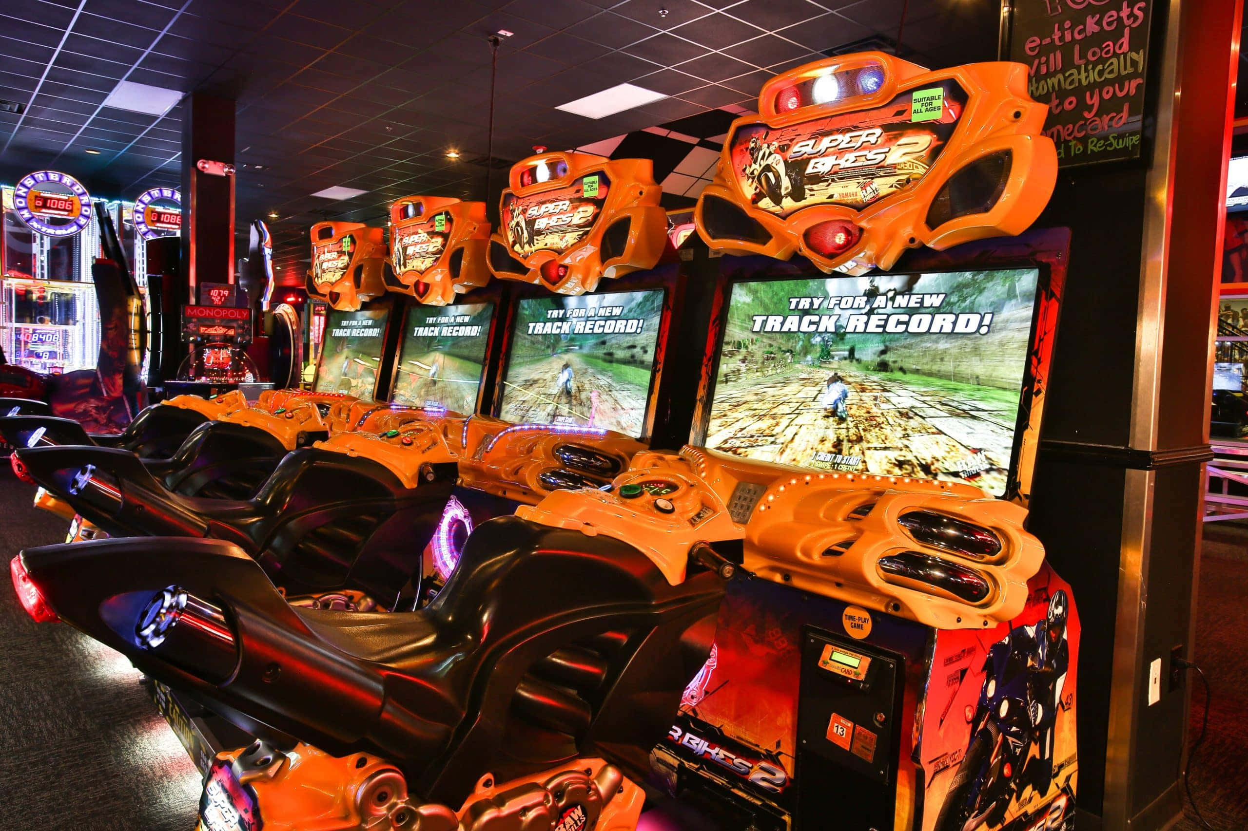 Arcade Games 桌布 壁紙 — 一排亮橙色賽車街機，配備發光屏幕和方向盤，位於燈光昏暗的遊戲廳。