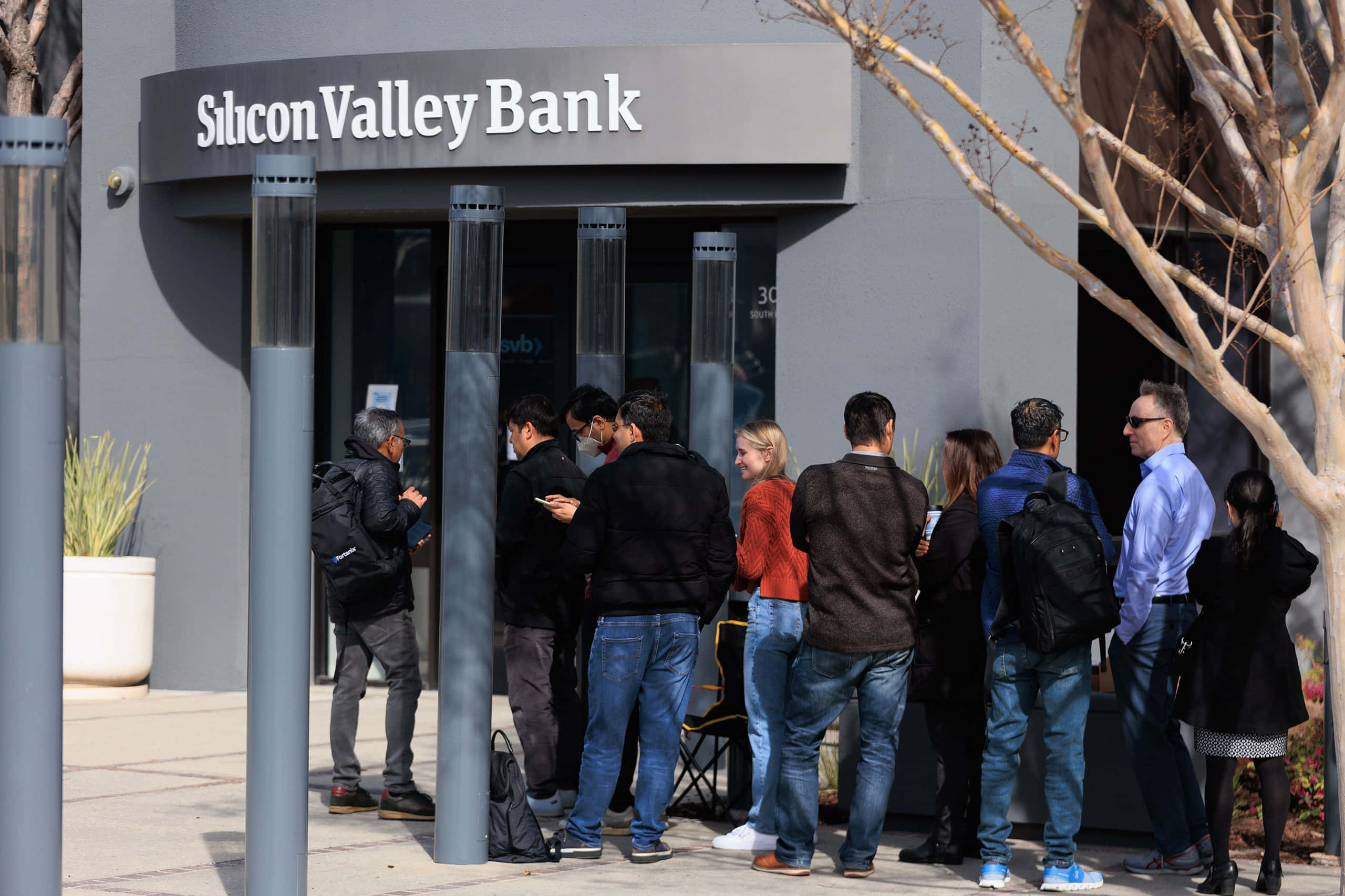 Bank 桌布 壁紙 — 一排人在陽光明媚的日子裡，在矽谷銀行分行外排隊等候，附近有光禿禿的樹木。