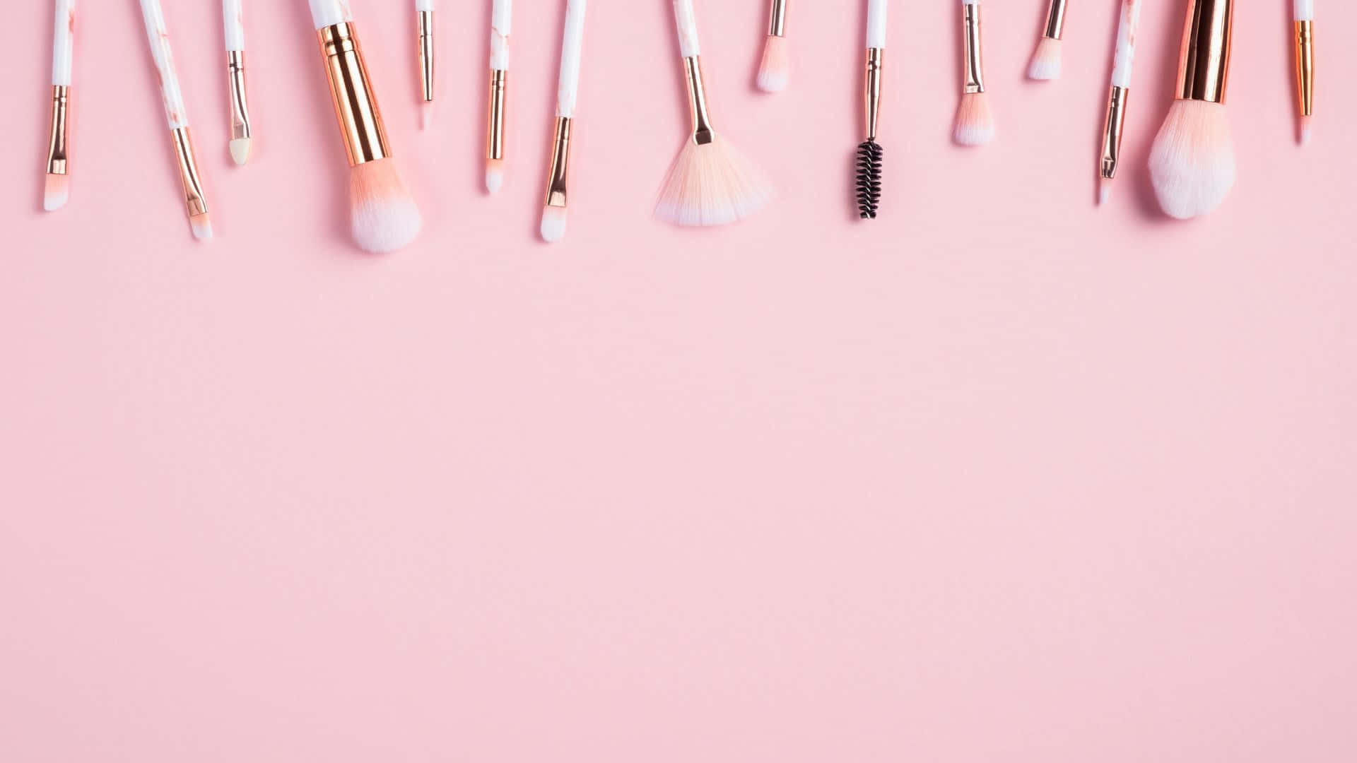 Pink Makeup 桌布 壁紙 — 一排各式各樣的化妝刷，帶有白色手柄和玫瑰金點綴，整齊地放置在柔和的粉紅色背景的頂部邊緣。