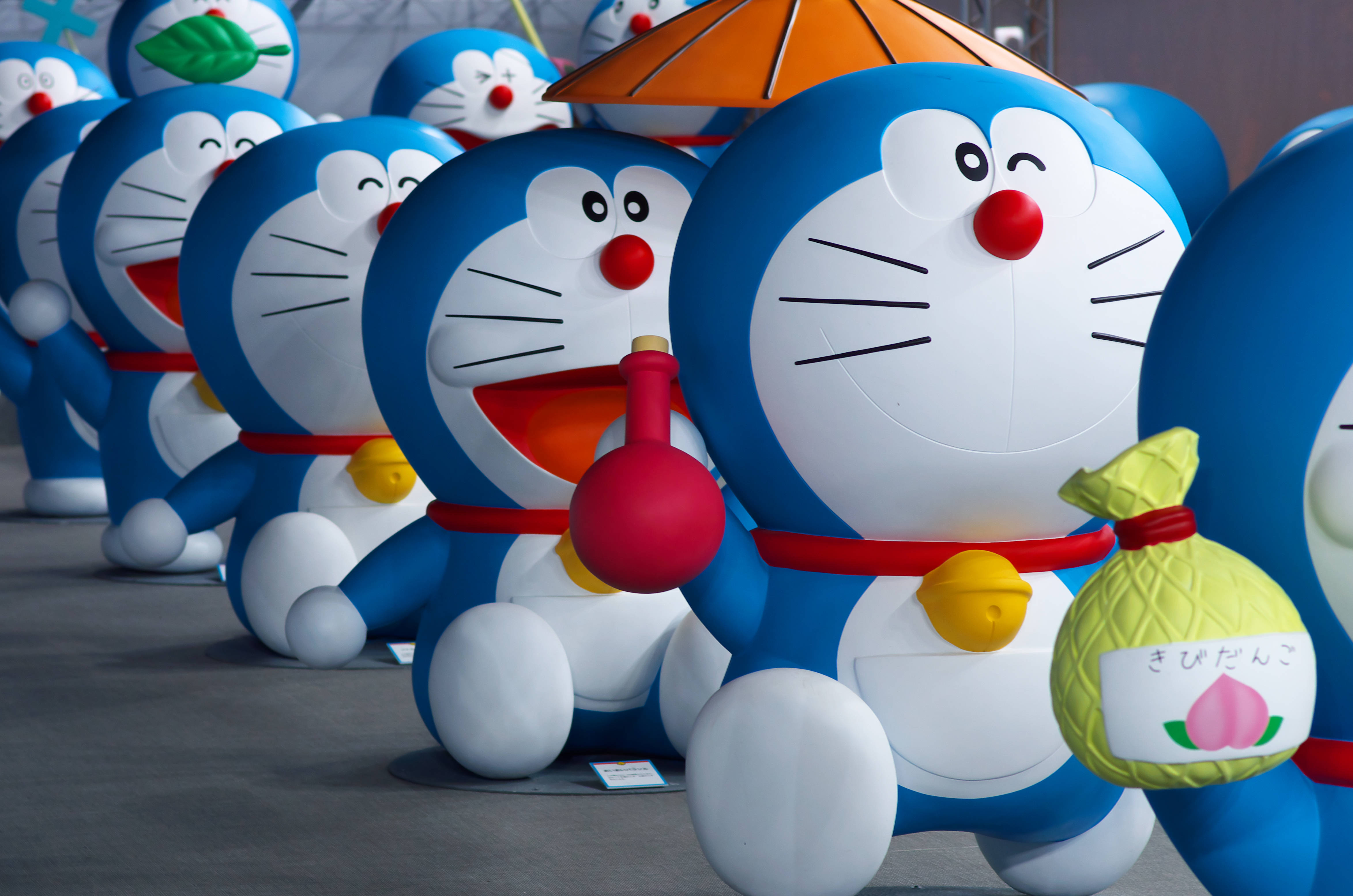 Doraemon 桌布 壁紙 — 一排大型藍色多啦A夢雕像手持紅色氣球和桃形袋，在公共展覽中愉快地微笑。