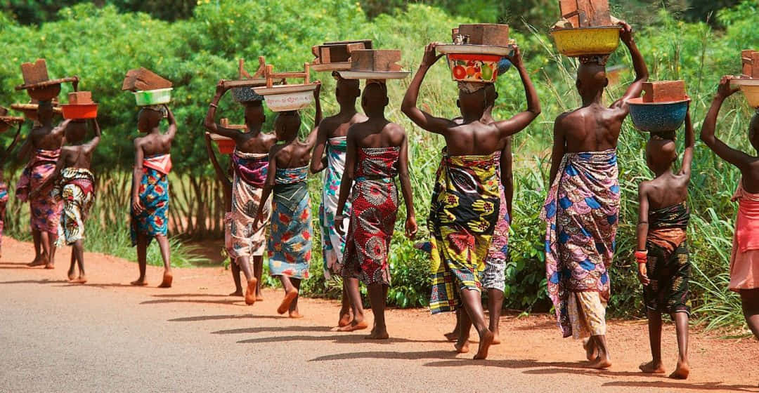 African Culture 桌布 壁紙 — 一排婦女和兒童穿著鮮豔圖案的衣服，赤腳沿著泥路行走，頭頂色彩繽紛的籃子，穿過茂密的綠植。