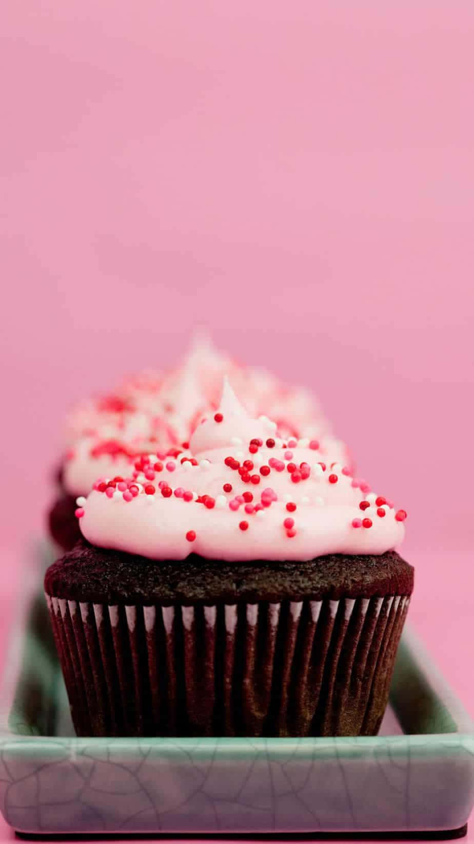 Cute Cupcake 桌布 壁紙 — 一排帶有粉紅色糖霜和紅色糖粒的朱古力杯子蛋糕，放在一個有裂紋的綠色碟子上，背景是柔和的粉紅色。