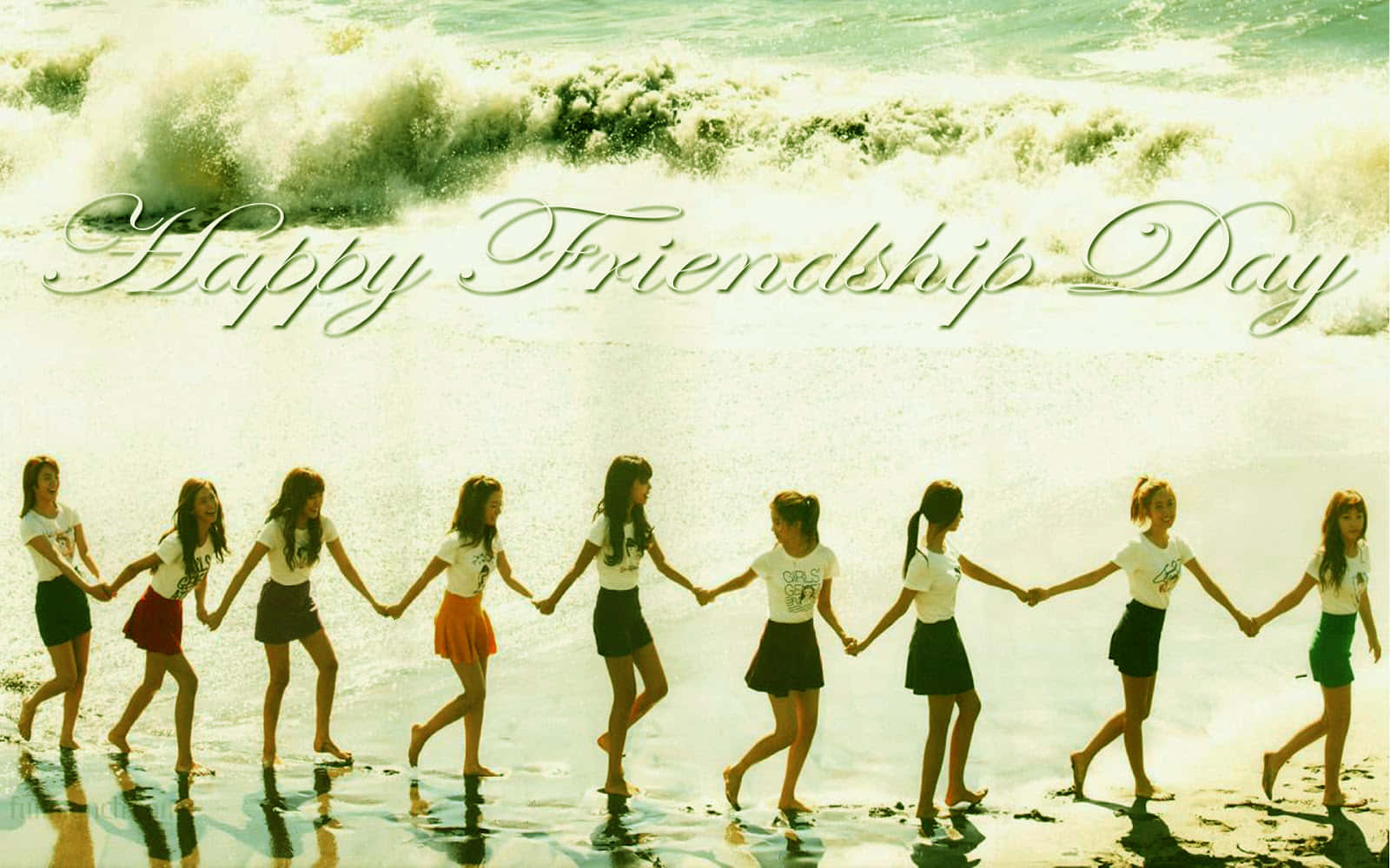 Friendship Day 桌布 壁紙 — 一排年輕女性手牽手沿著陽光明媚的海灘行走，身後海浪拍打，上方寫有「Happy Friendship Day」字樣。