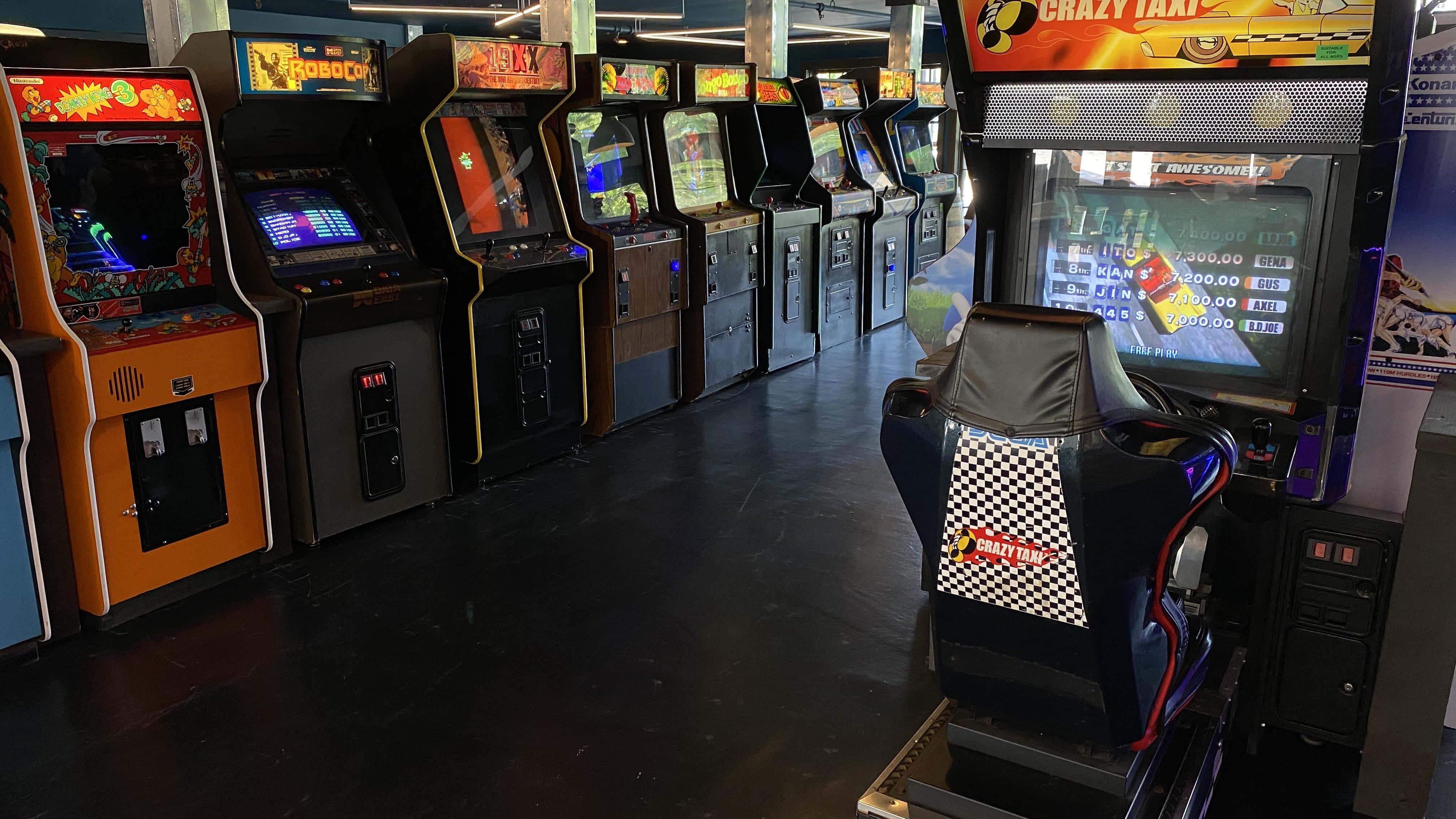 Arcade Games 桌布 壁紙 — 一排復古街機，帶有發光的屏幕和色彩繽紛的機櫃，排列在燈光昏暗的遊戲廳中。