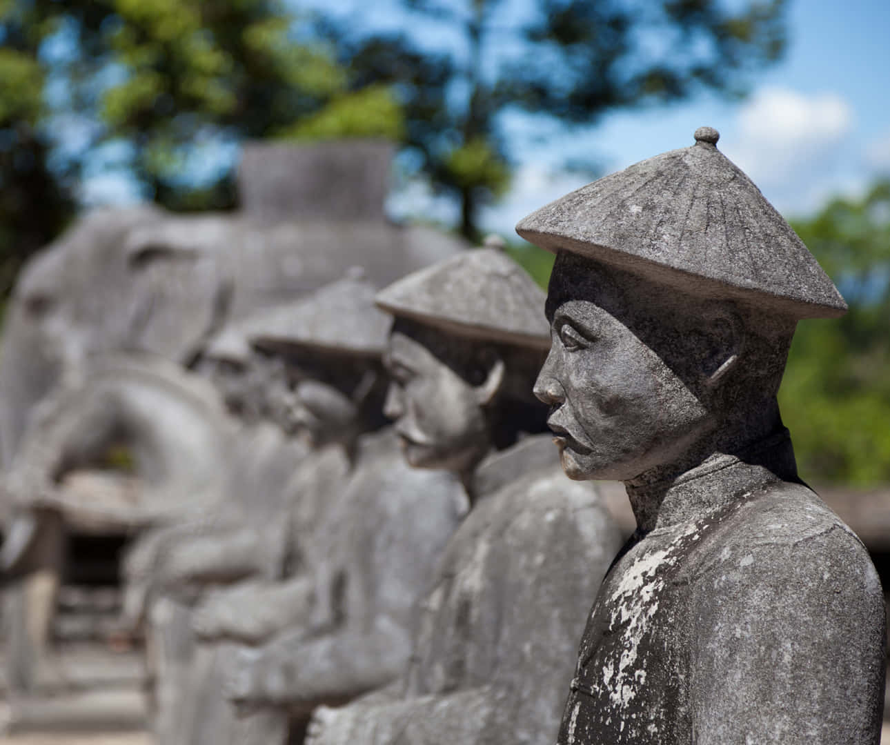Vietnam 桌布 壁紙 — 一排戴著傳統錐形帽的男性風化石像，在明亮的藍天下，以綠樹為背景，莊嚴地矗立著。