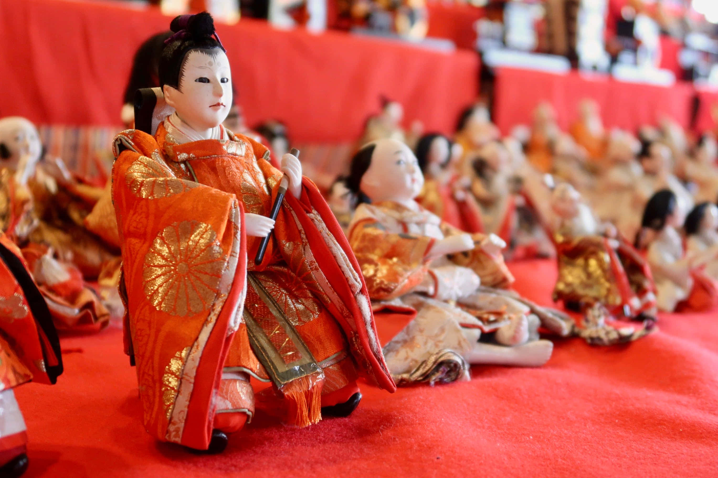 Dolls 桌布 壁紙 — 一排排傳統日本雛人形穿著華麗的橙色和金色和服，優雅地坐在鮮紅色的地毯上。