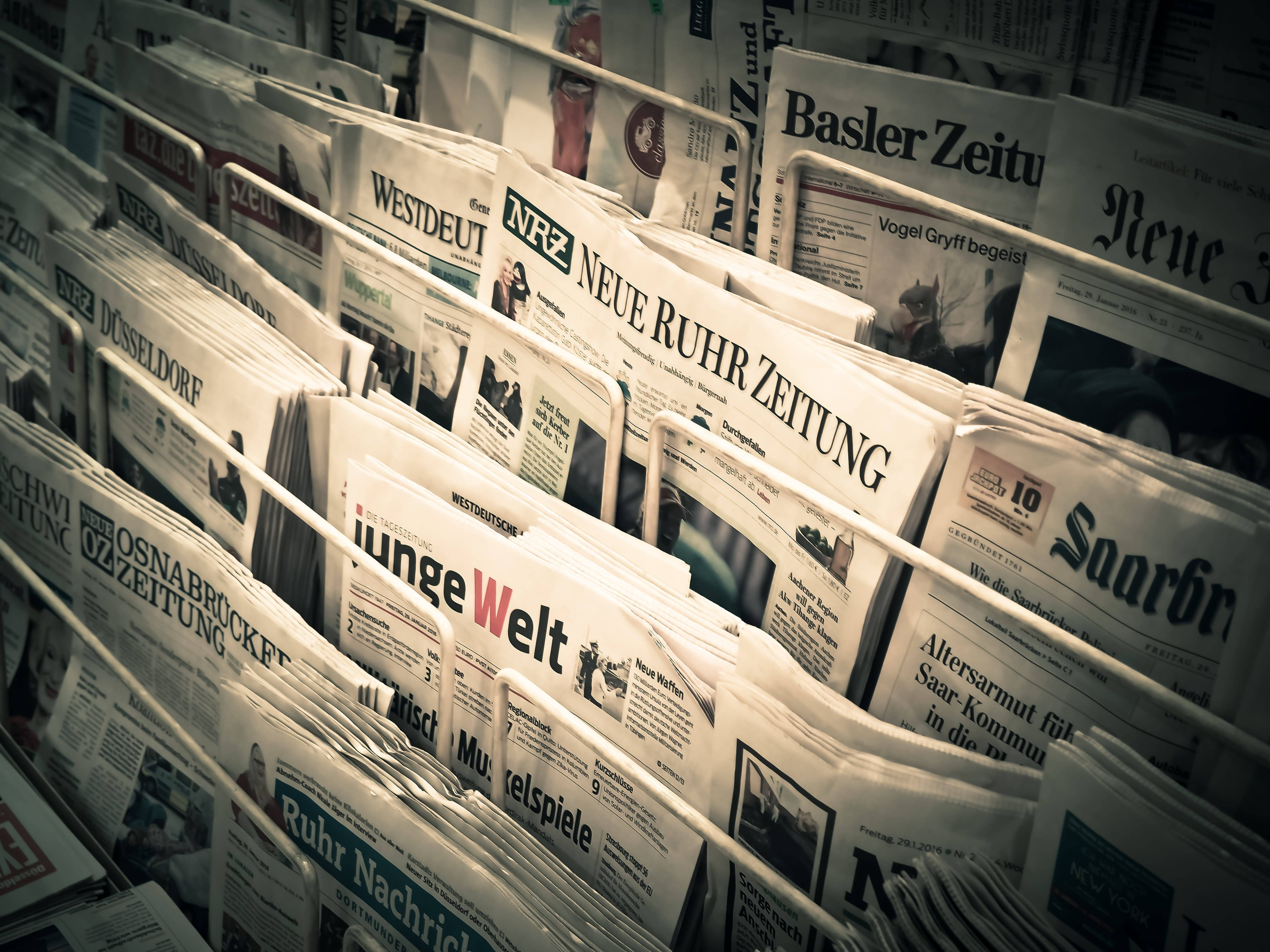 Newspaper 桌布 壁紙 — 一排排德國報紙放在架上，暖色燈光下標題清晰可見。