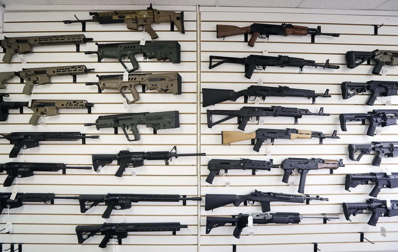 槍 桌布 壁紙 — 一排排步槍和突擊武器陳列在槍店的白色板條牆上，配有螢光燈照明。