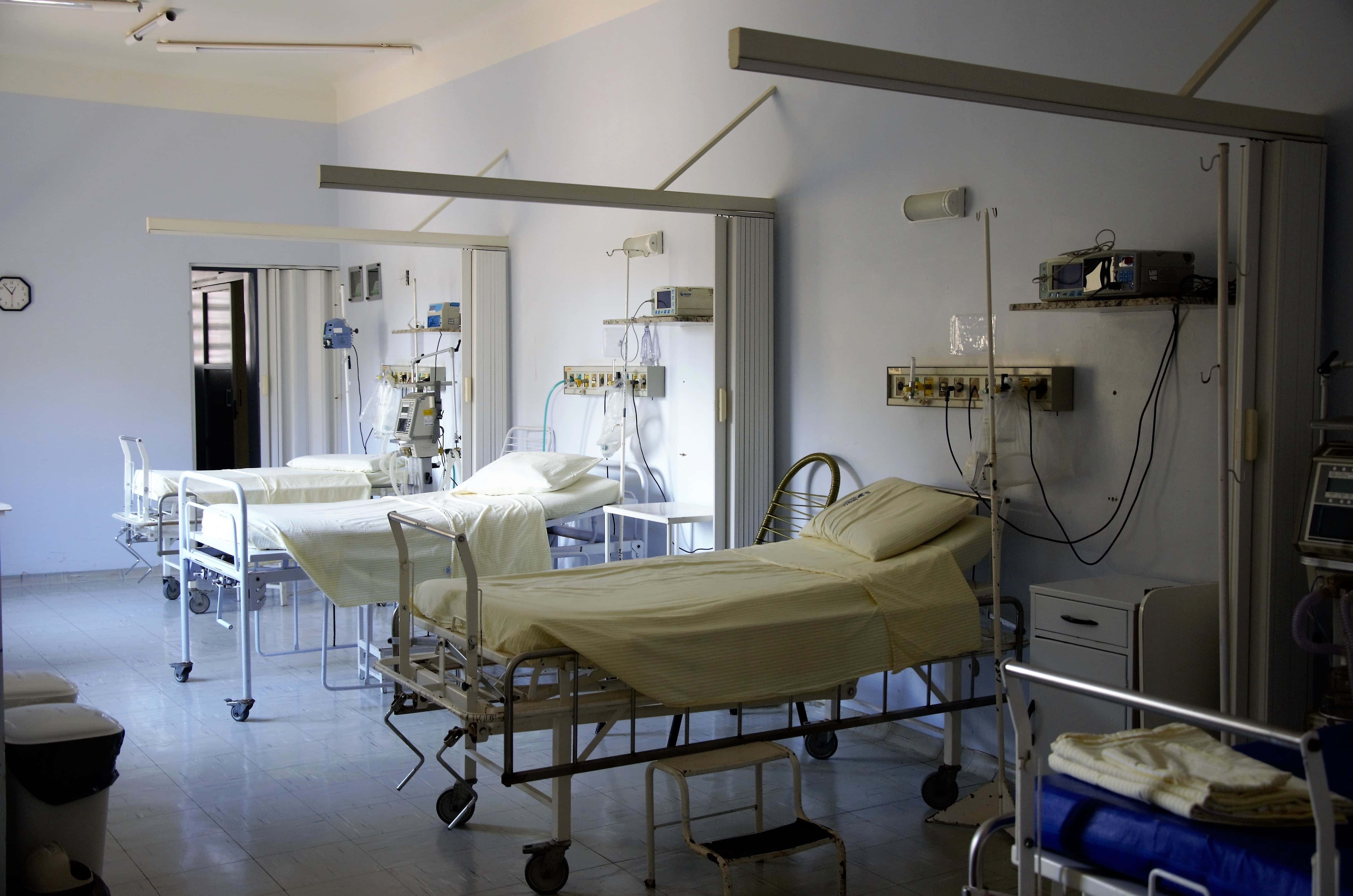 Hospital Bed 桌布 壁紙 — 一排排空置的病床連同醫療設備在螢光燈下排列在無菌的白色病房中。