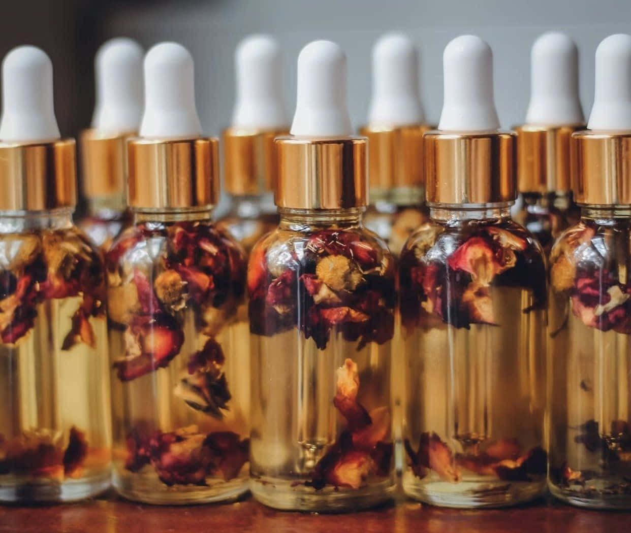 Rose Oil 桌布 壁紙 — 一排排裝有金色液體和乾燥玫瑰花瓣的玻璃滴管瓶放在木質表面上。