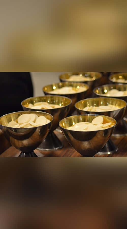 Sacrament 桌布 壁紙 — 一排排閃閃發光的金色聖杯碗，裝滿了淺色圓形薄餅，排列在木質表面上。