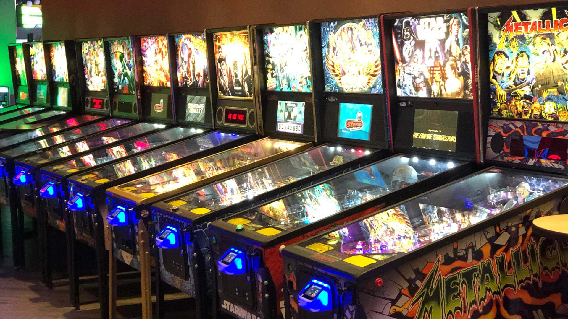 Arcade Games 桌布 壁紙 — 一排明亮照明的復古彈珠機，帶有彩色藝術品和發光顯示屏，位於復古遊戲機中心。