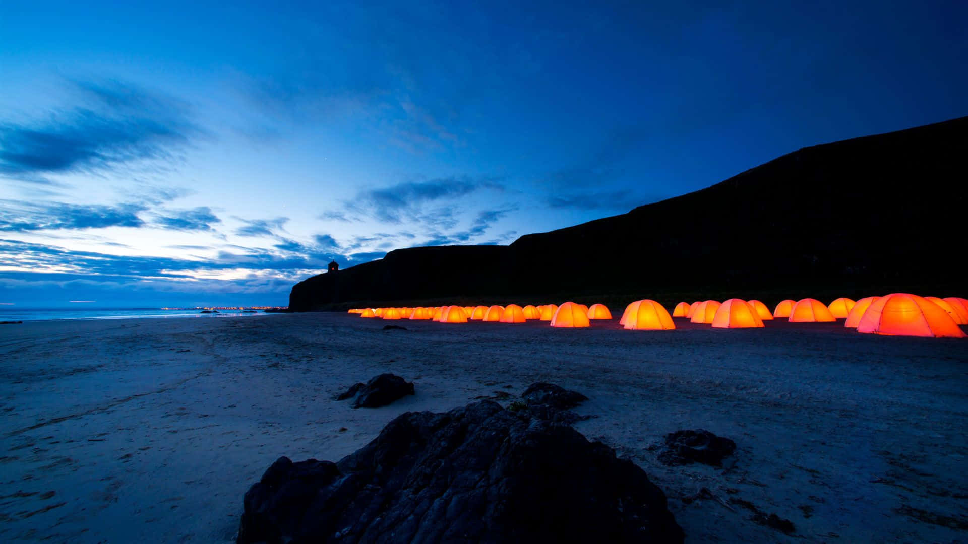 Beach Camping 桌布 壁紙 — 一排發光的橙色帳篷排列在暮色中寧靜的海灘上，背景是黑暗的懸崖和深藍色的天空。
