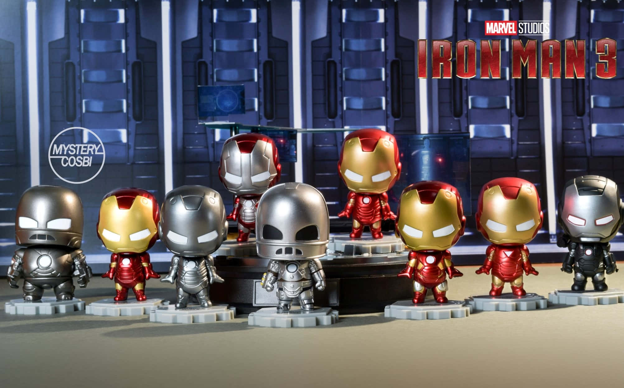 Iron Man Bobbleheads 桌布 壁紙 — 一排穿著不同裝甲服的Q版鐵甲奇俠人偶，在帶有『鐵甲奇俠3』品牌標誌的科技主題舞台上展示。