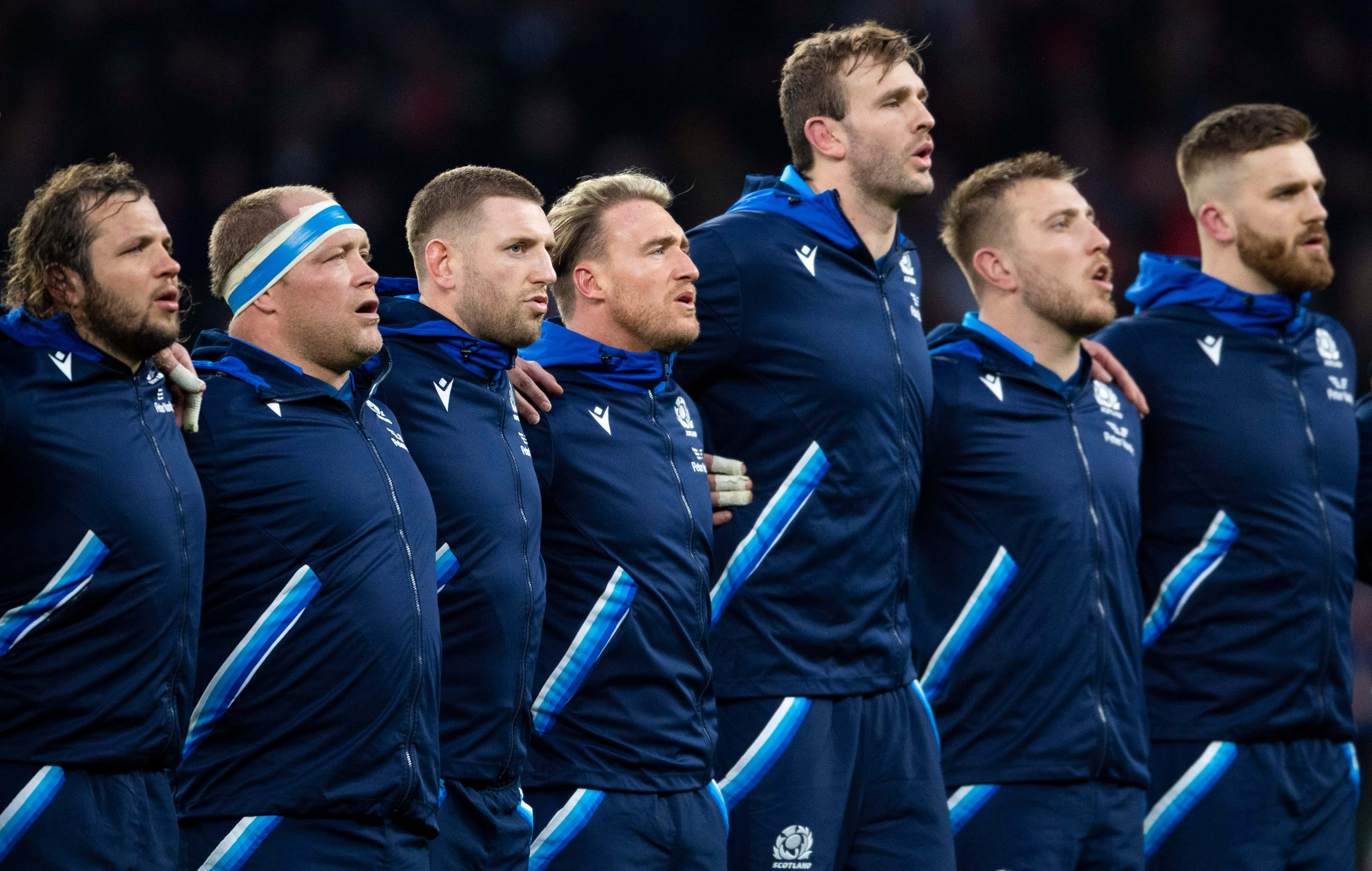 Scotland Rugby 桌布 壁紙 — 一排穿著藍色運動服的欖球運動員在國歌演奏期間莊嚴地站著，手臂互相搭在對方肩上。