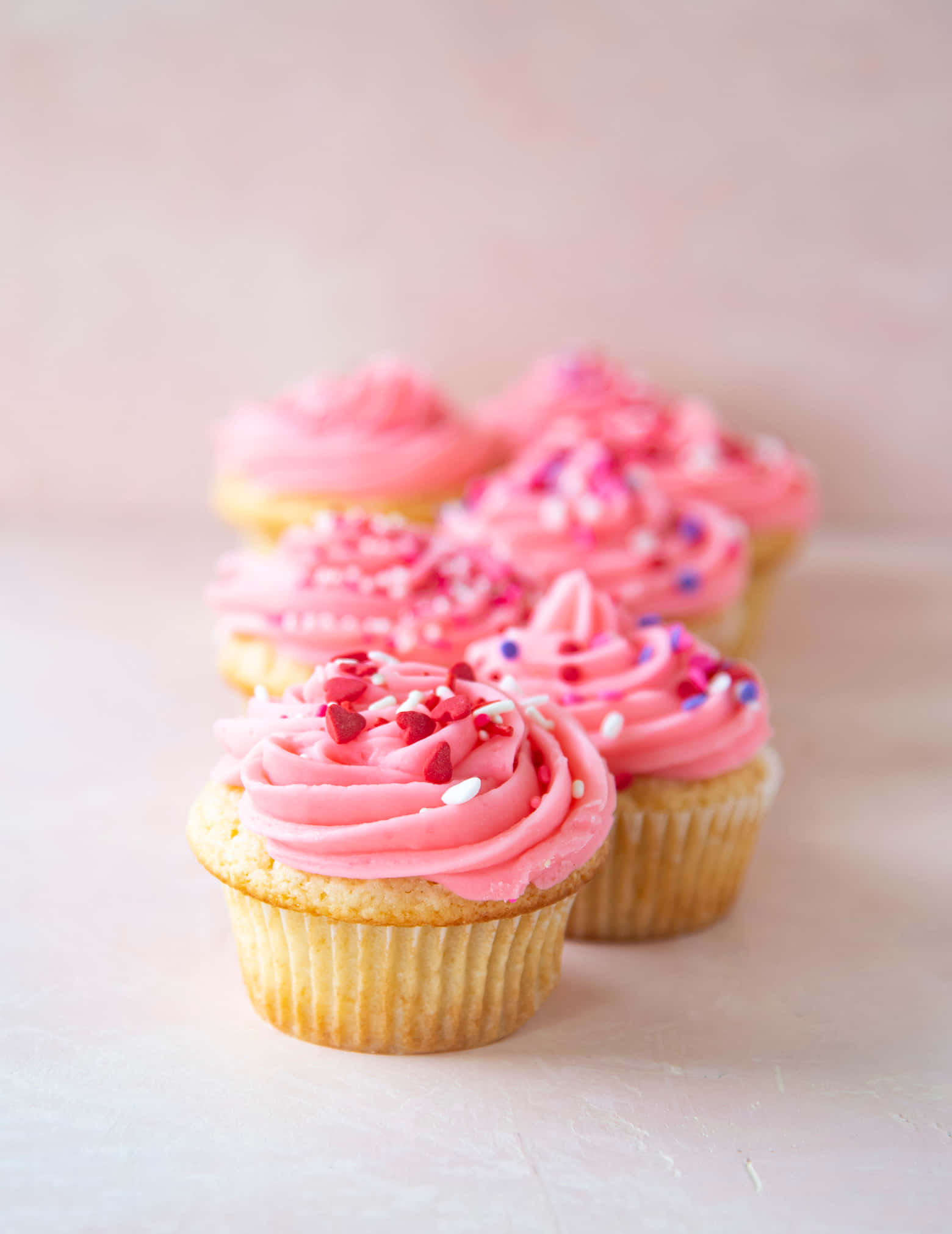 Pink Cupcakes 桌布 壁紙 — 一排粉紅色糖霜的杯子蛋糕，上面撒有心形裝飾，放在柔軟的粉紅色表面上，以淺景深誘人地排列。