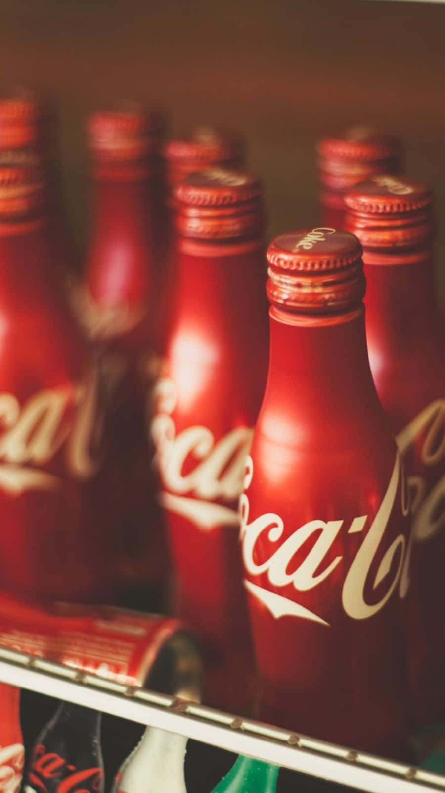 coca cola 桌布 壁紙 — 一排經典的紅色可口可樂玻璃瓶，帶有白色字樣，整齊地放在雪櫃架上，柔和地以暖色調照明。