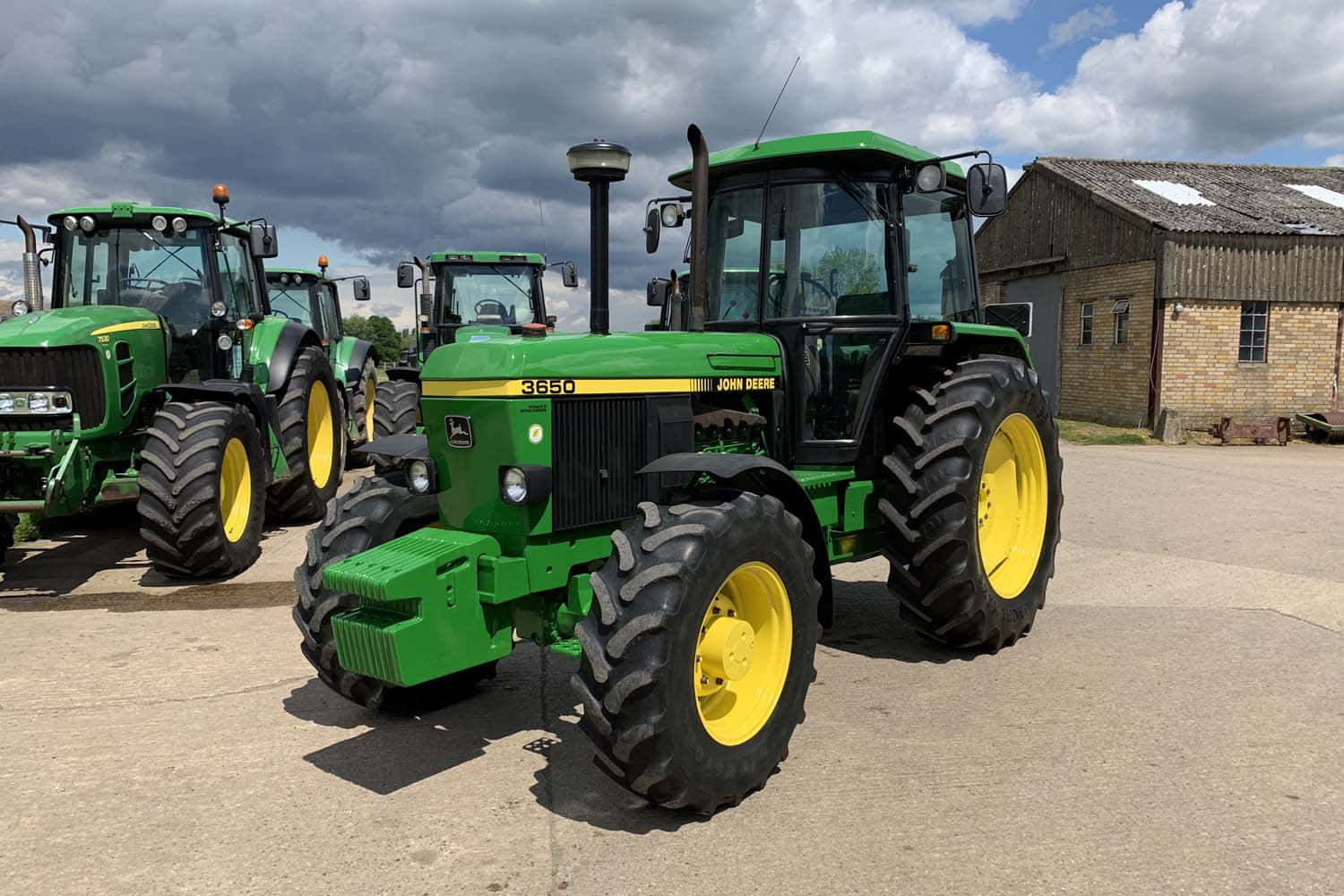 John Deere 桌布 壁紙 — 一排綠色和黃色的約翰迪爾拖拉機停泊在混凝土上，天空多雲，靠近一座鄉村穀倉。