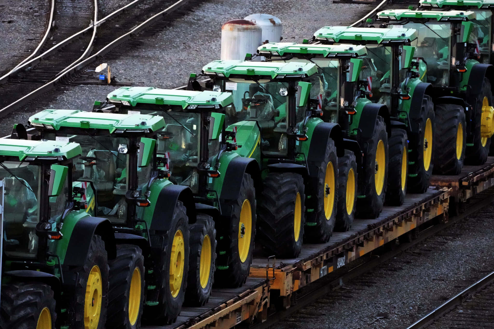 John Deere 桌布 壁紙 — 一排綠色約翰迪爾拖拉機，黃色車輪，整齊地排列在碎石場的平板火車車廂上。